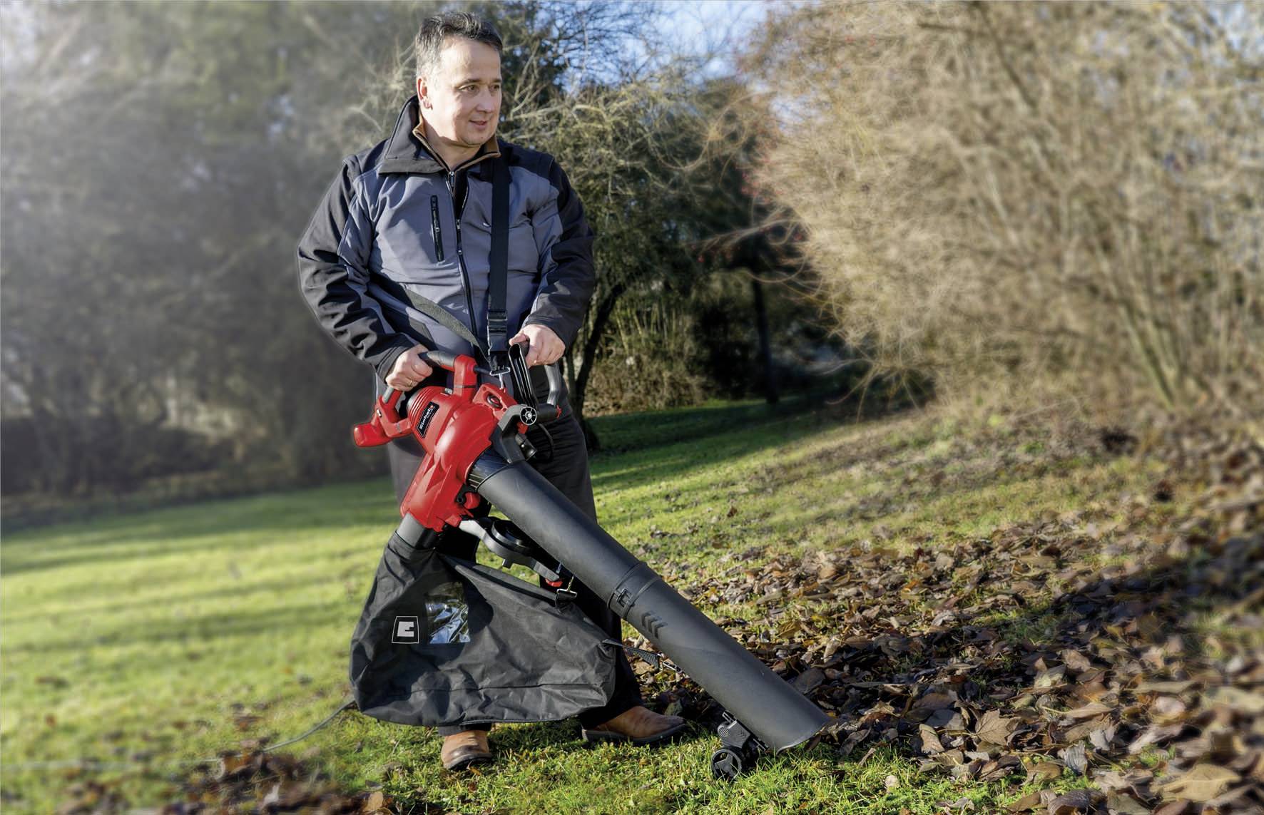 Muž používá červený zahradní vysavač na trávníku, aby odstranil listí. Má na sobě bundu a stojí vedle stromů v pozadí.