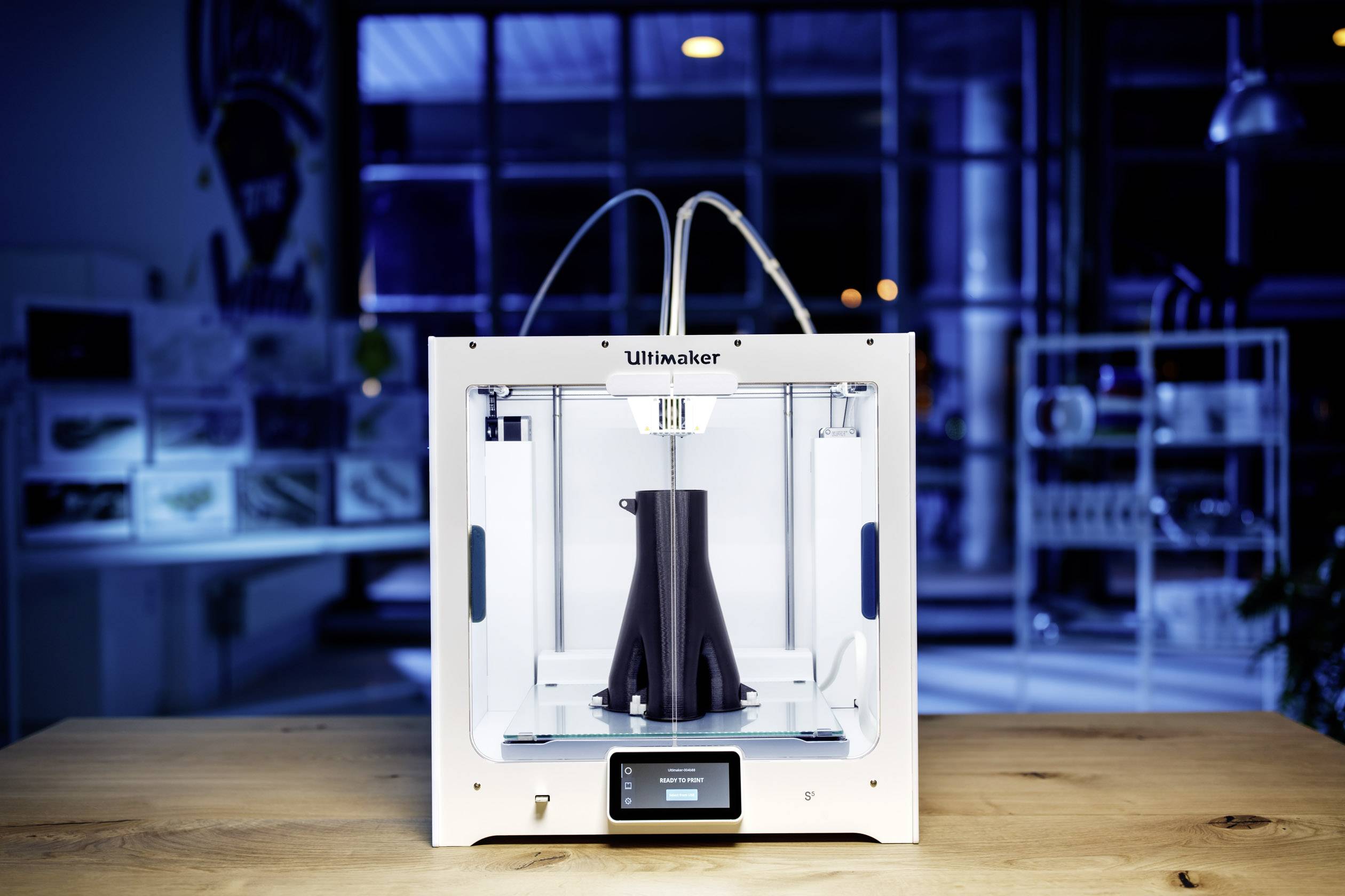 3D tiskárna tiskne černý objekt v moderním prostředí dílny. Tiskárna má bílou skříň a displej.