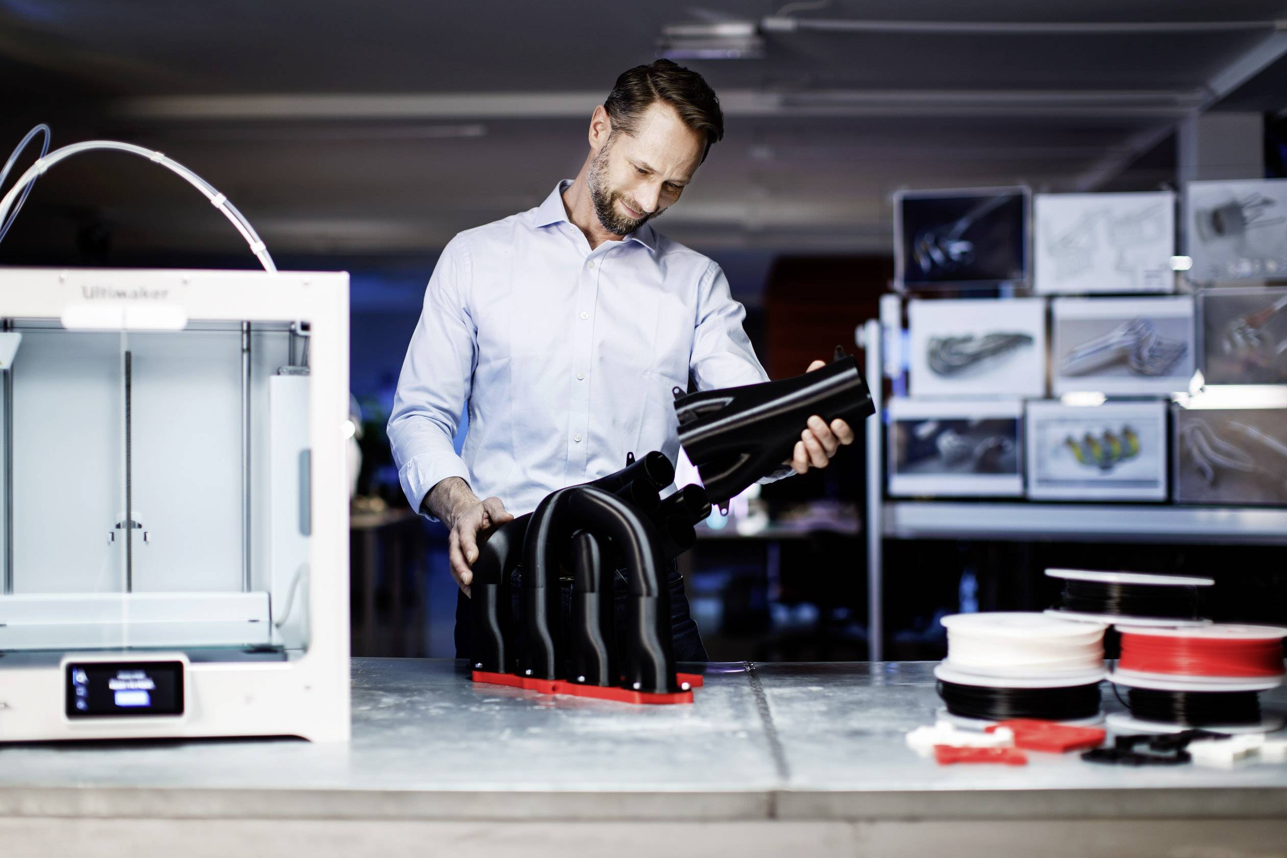 Muž pozoruje velký 3D tištěný objekt v moderní laboratoři. Vedle něj stojí 3D tiskárna a různé druhy filamentů.