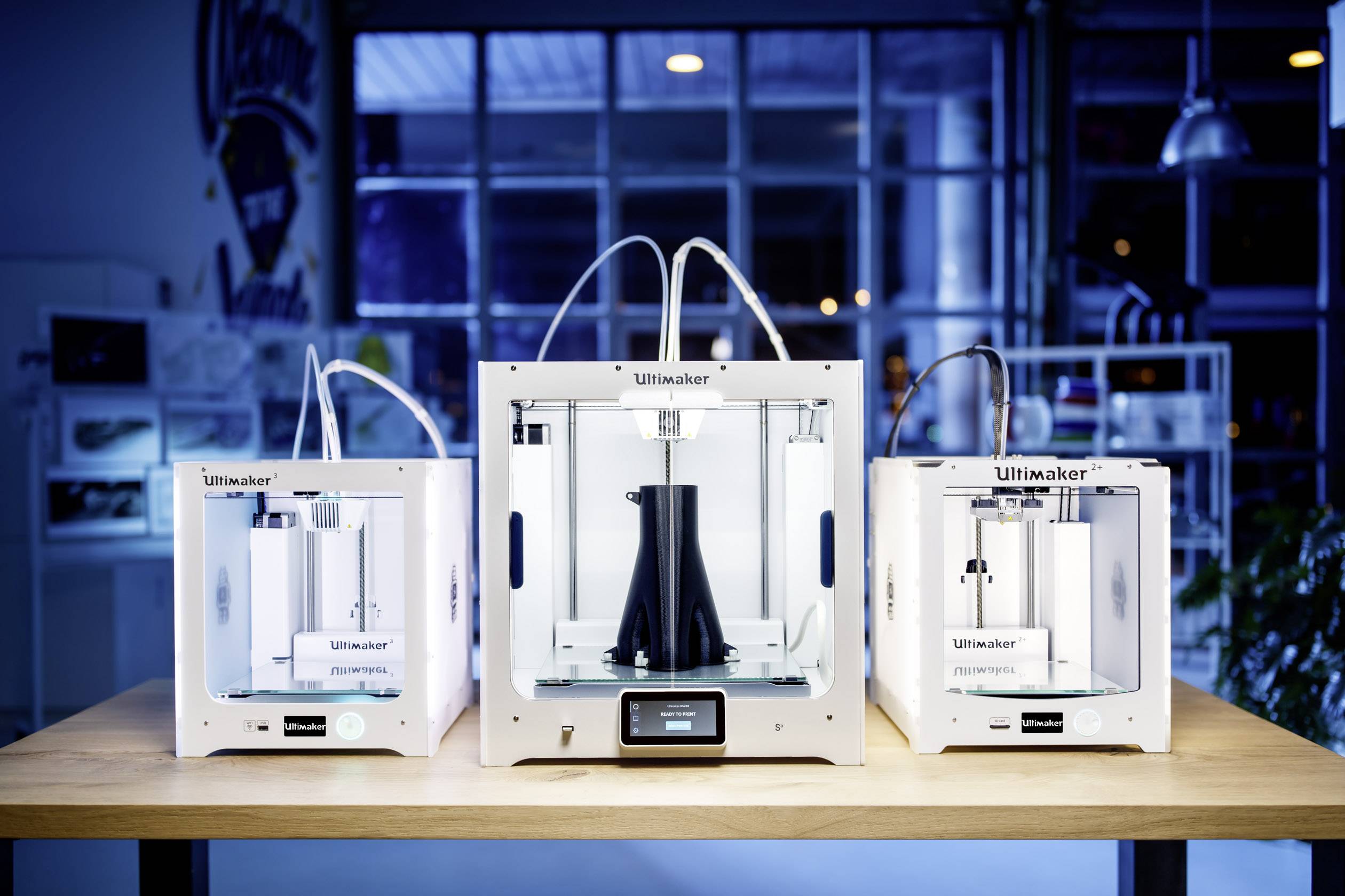 Tři 3D tiskárny značky Ultimaker stojí na stole. Prostřední tiskárna je větší a obsahuje černý, kuželovitý objekt.