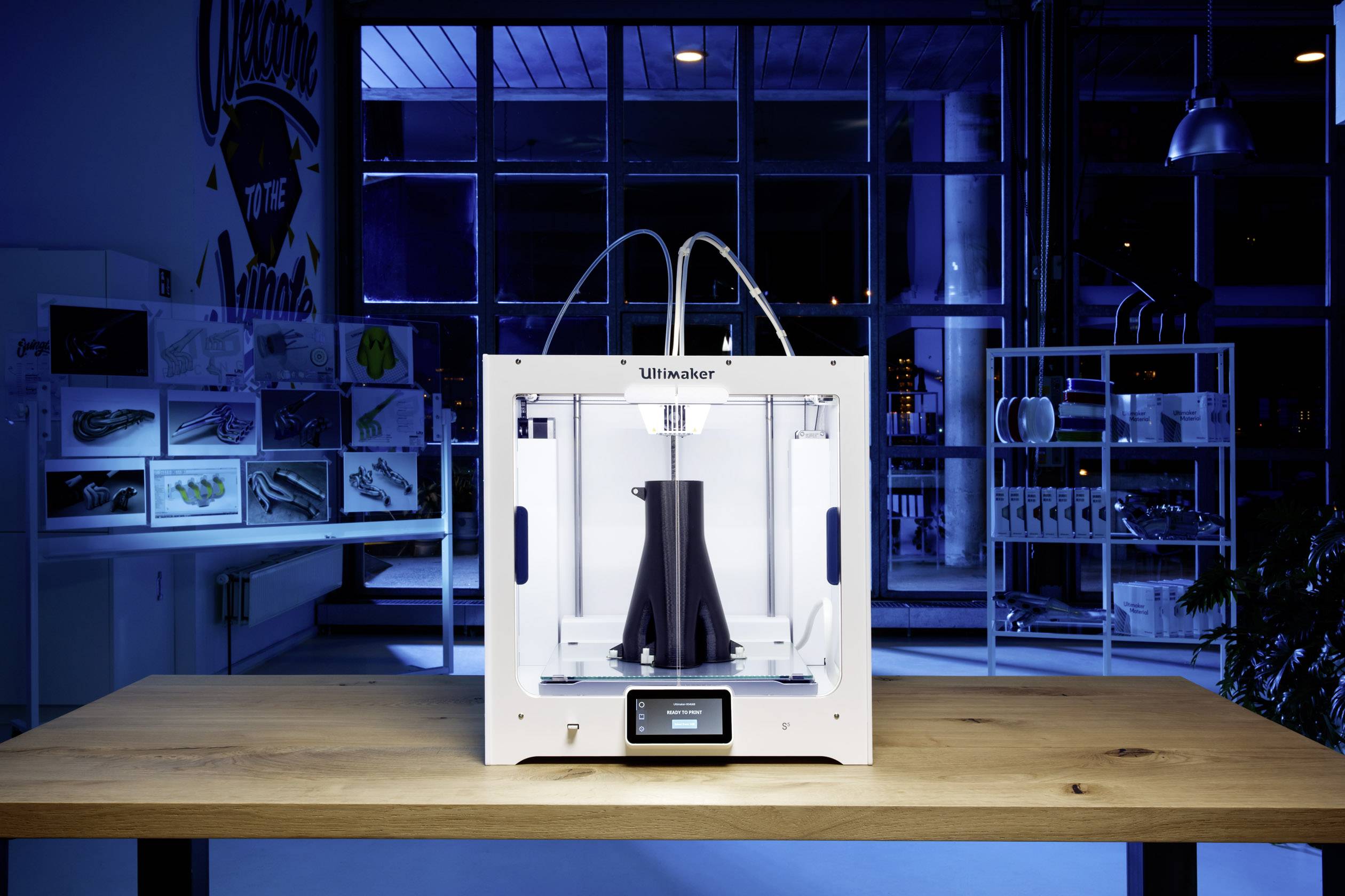 Moderní 3D tiskárna umístěná na stole tiskne černý objekt. V pozadí jsou viditelné police s modelovými prototypy.