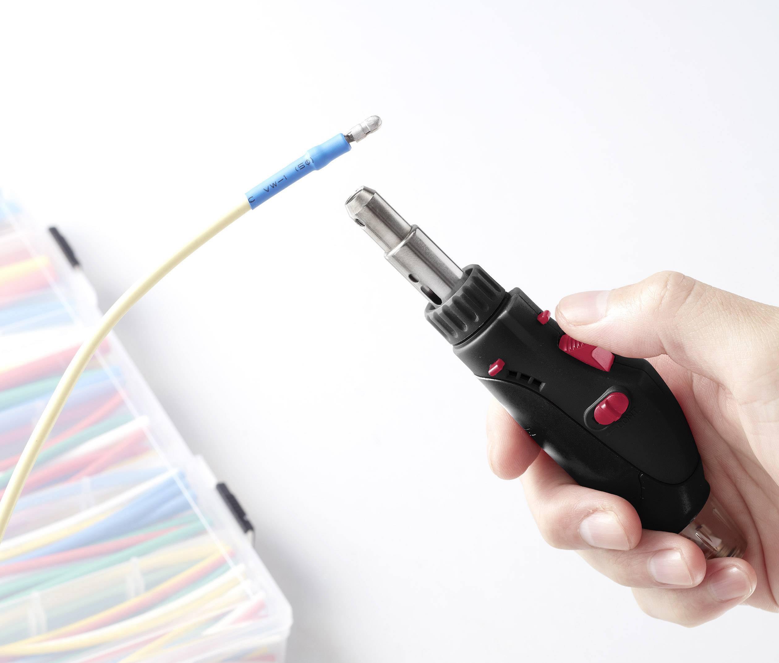 Jedna ruka drží plynový hořák namířený na žlutý kabel s modrou plastovou izolací; v pozadí je organizér s barevnými kabely.