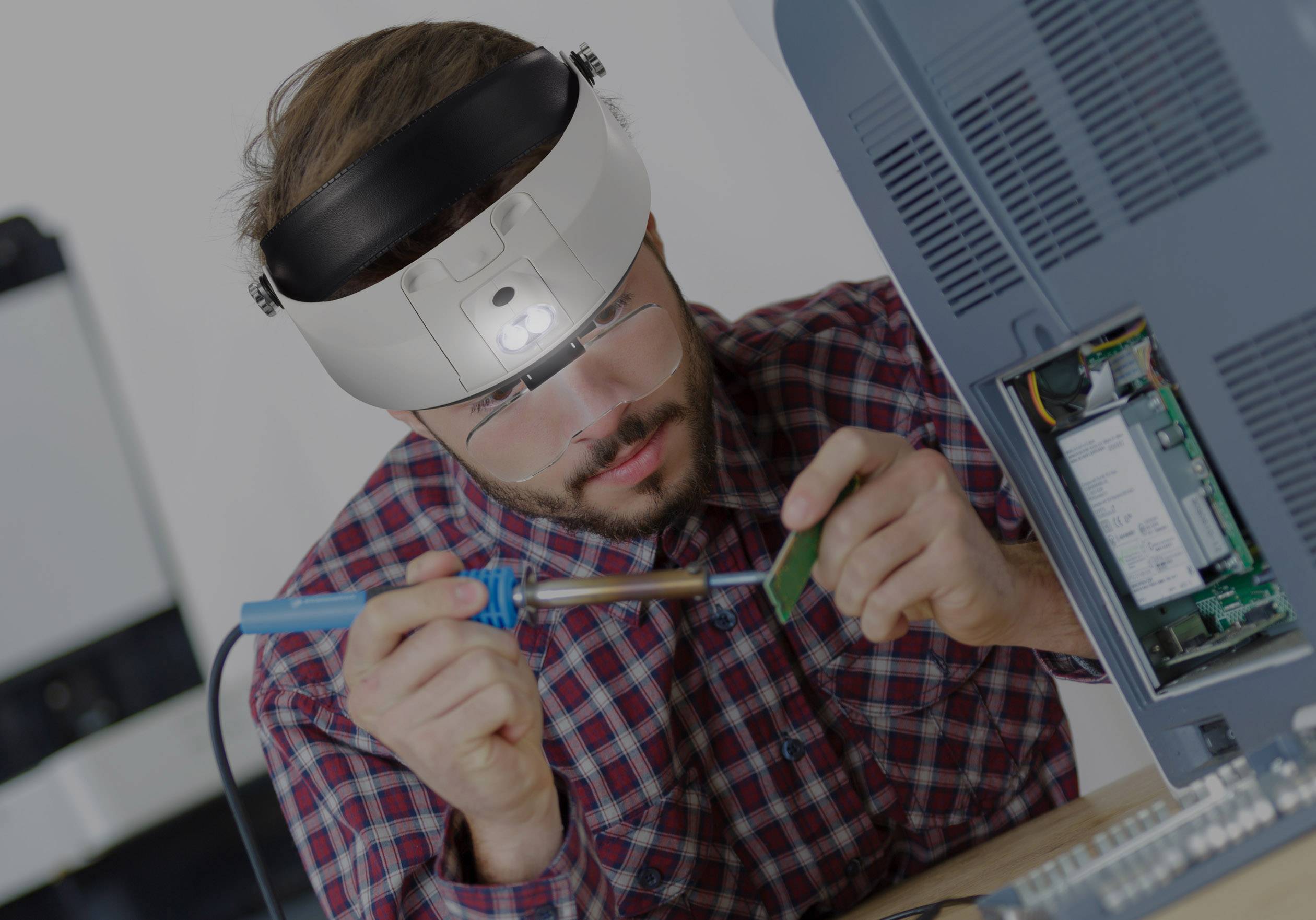 Muž soustředěně pájí na elektronické desce zařízení. Má nasazené lupové brýle pro přesnější práci.