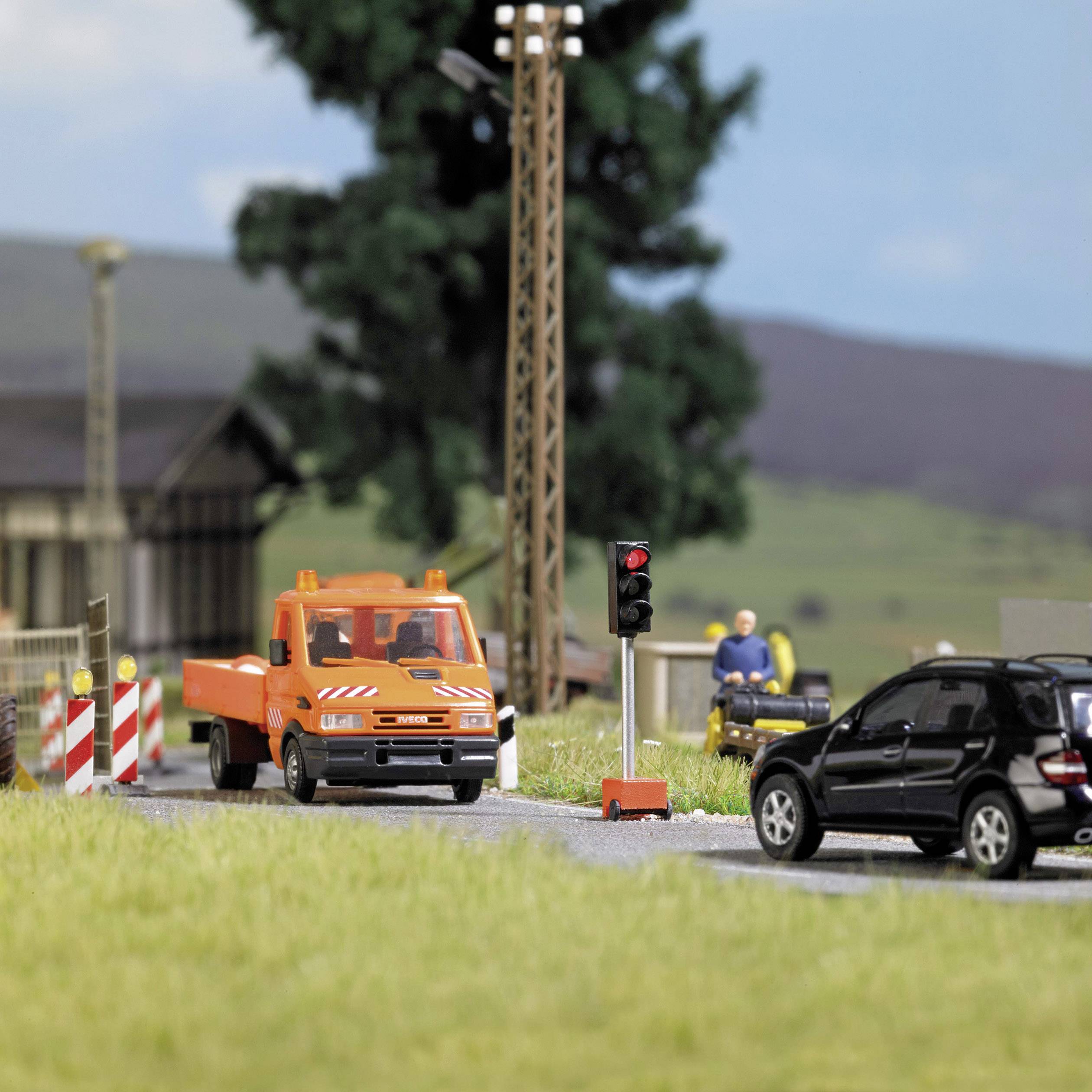 Oranžový nákladní vůz stojí na staveništi u silnice, zatímco černé auto čeká na semaforu. V pozadí jsou stromy.