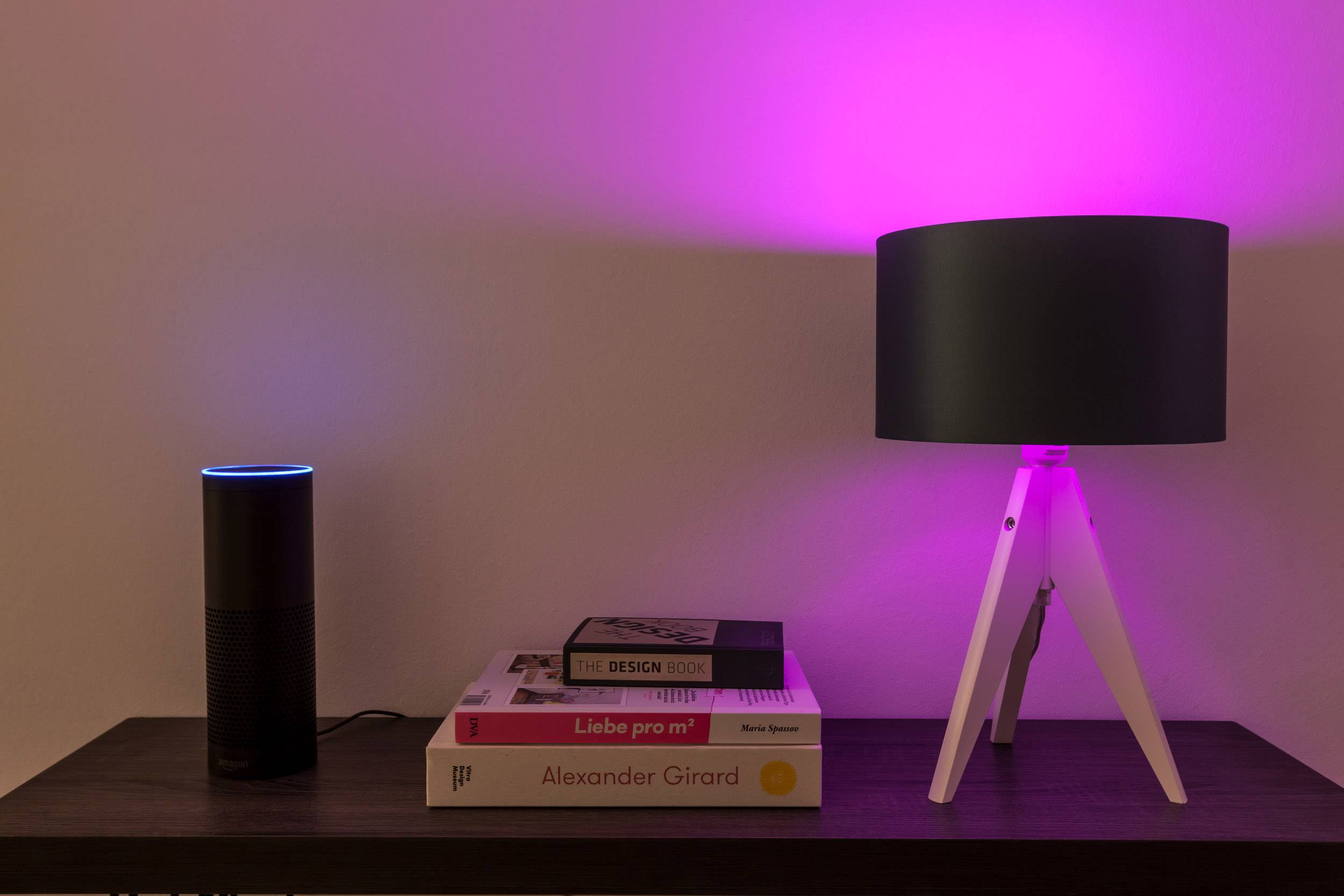 Chytrý reproduktor a stolní lampa svítící růžově stojí na stole vedle stohu knih.