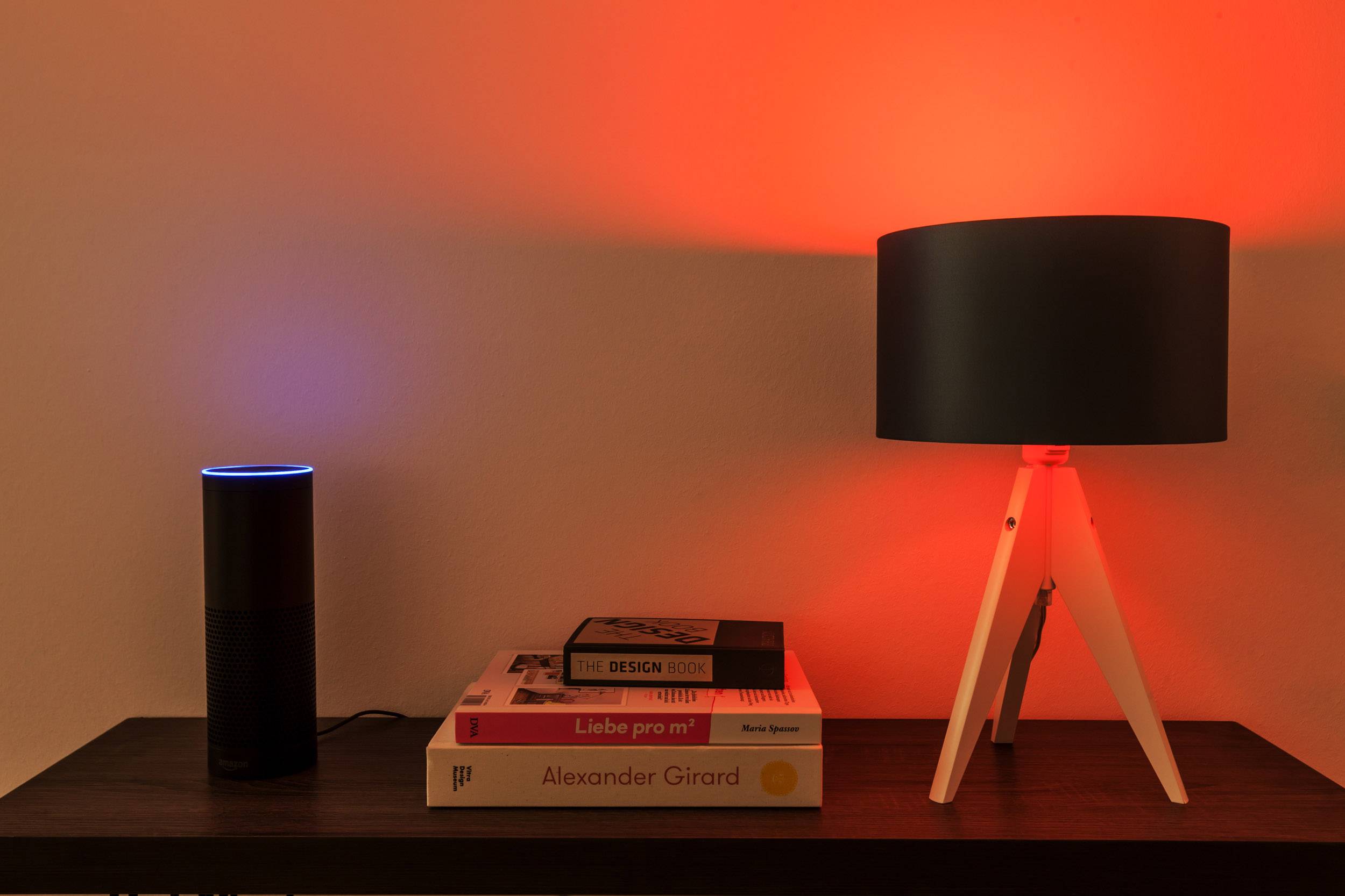 Chytrý reproduktor se svítícím modrým kroužkem stojí vedle stohu knih a osvětlené stolní lampy na stole.