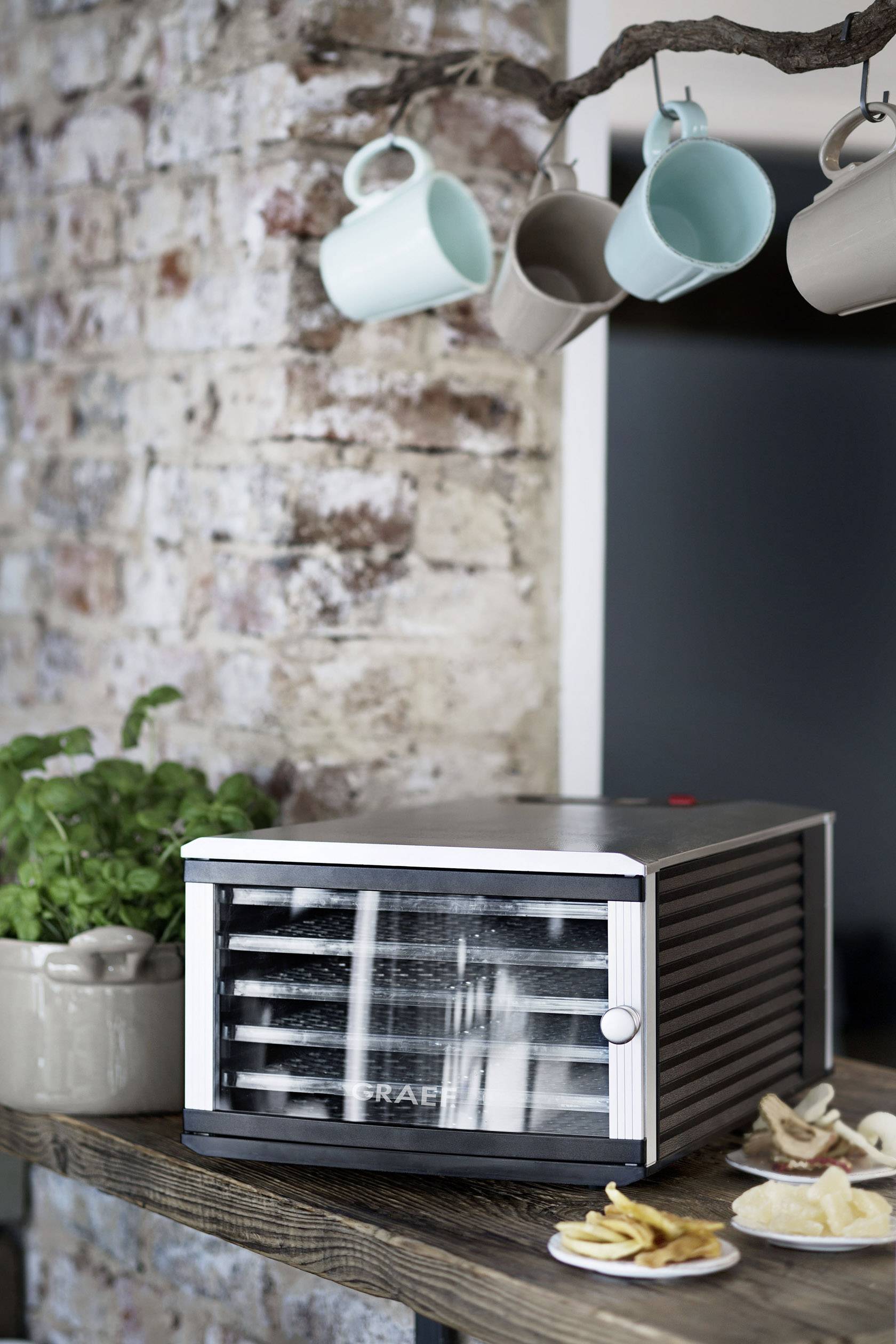 Odšťavňovač na rustikálním dřevěném stole vedle sušeného ovoce a bylinkového hrnku, obklopený visícími šálky před cihlovou zdí.