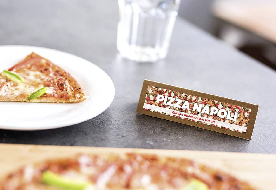Kousek pizzy na talíři vedle cedule s nápisem 'Pizza Napoli'. V pozadí stojí sklenice vody.