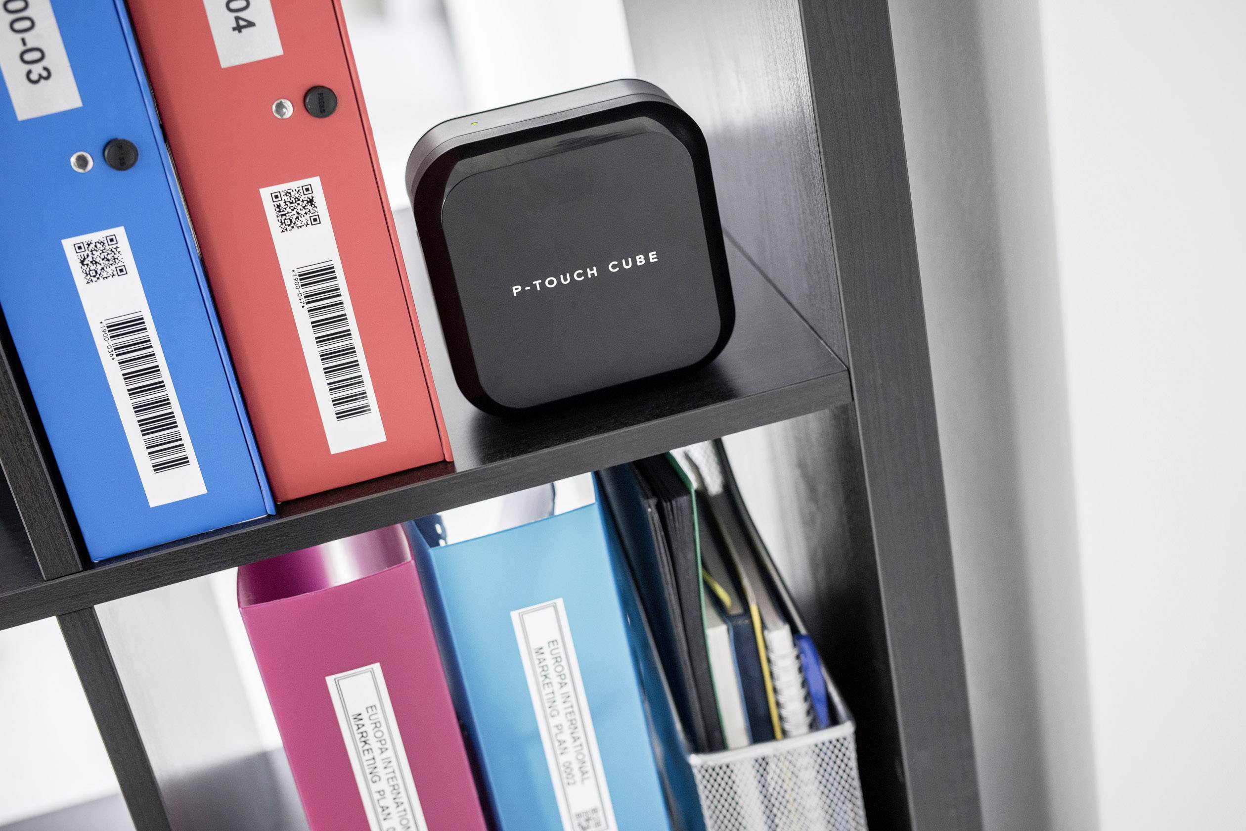 Černé čtvercové zařízení s nápisem 'P-Touch Cube' stojí na polici. Vedle něj jsou barevné šanony.