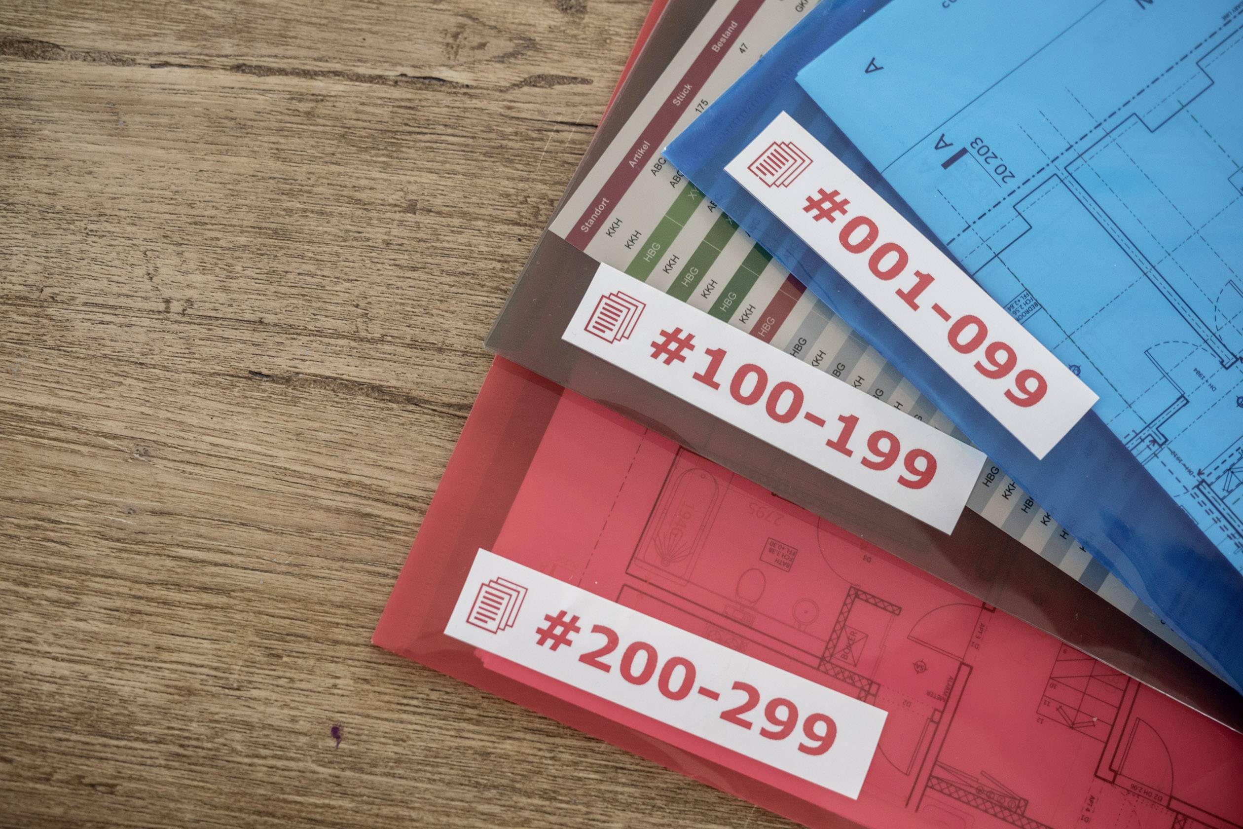 Tři složky s plány leží na dřevěném stole. Jsou označeny čísly '#001-099', '#100-199' a '#200-299'.