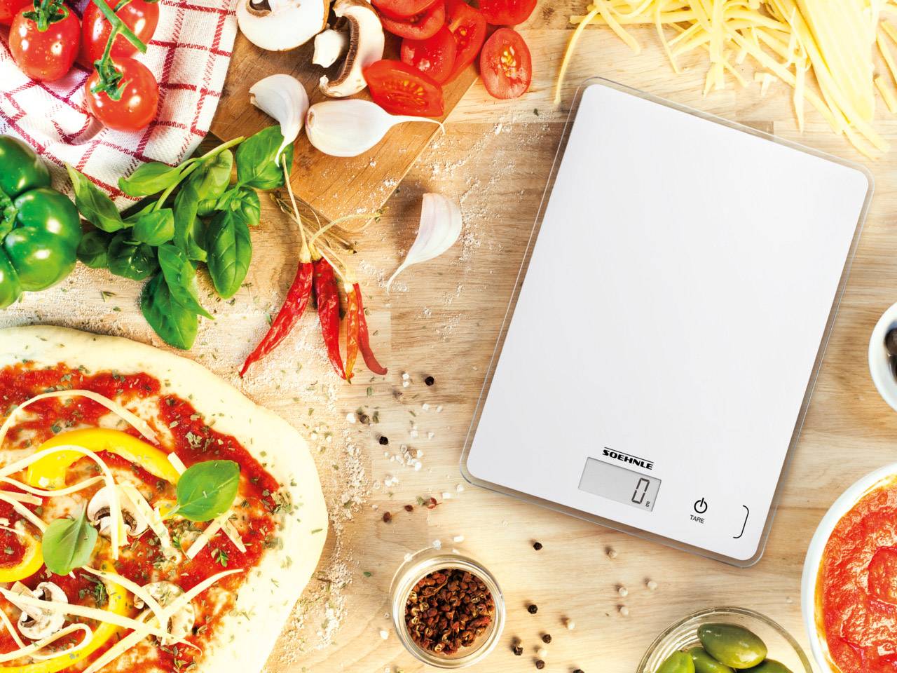 Digitální kuchyňská váha na dřevěném stole, obklopená čerstvými ingrediencemi na pizzu, jako jsou těsto, zelenina, bylinky a koření. Displej ukazuje '0'.