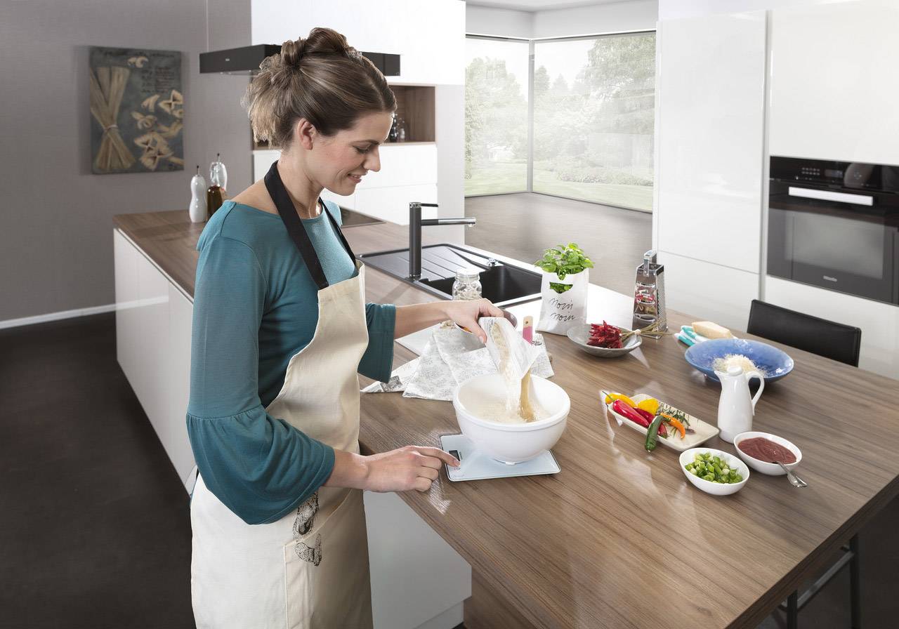 Žena peče v moderní kuchyni. Drží lžíci a míchá ve velké míse. Ingredience jsou rozmístěné na stole.