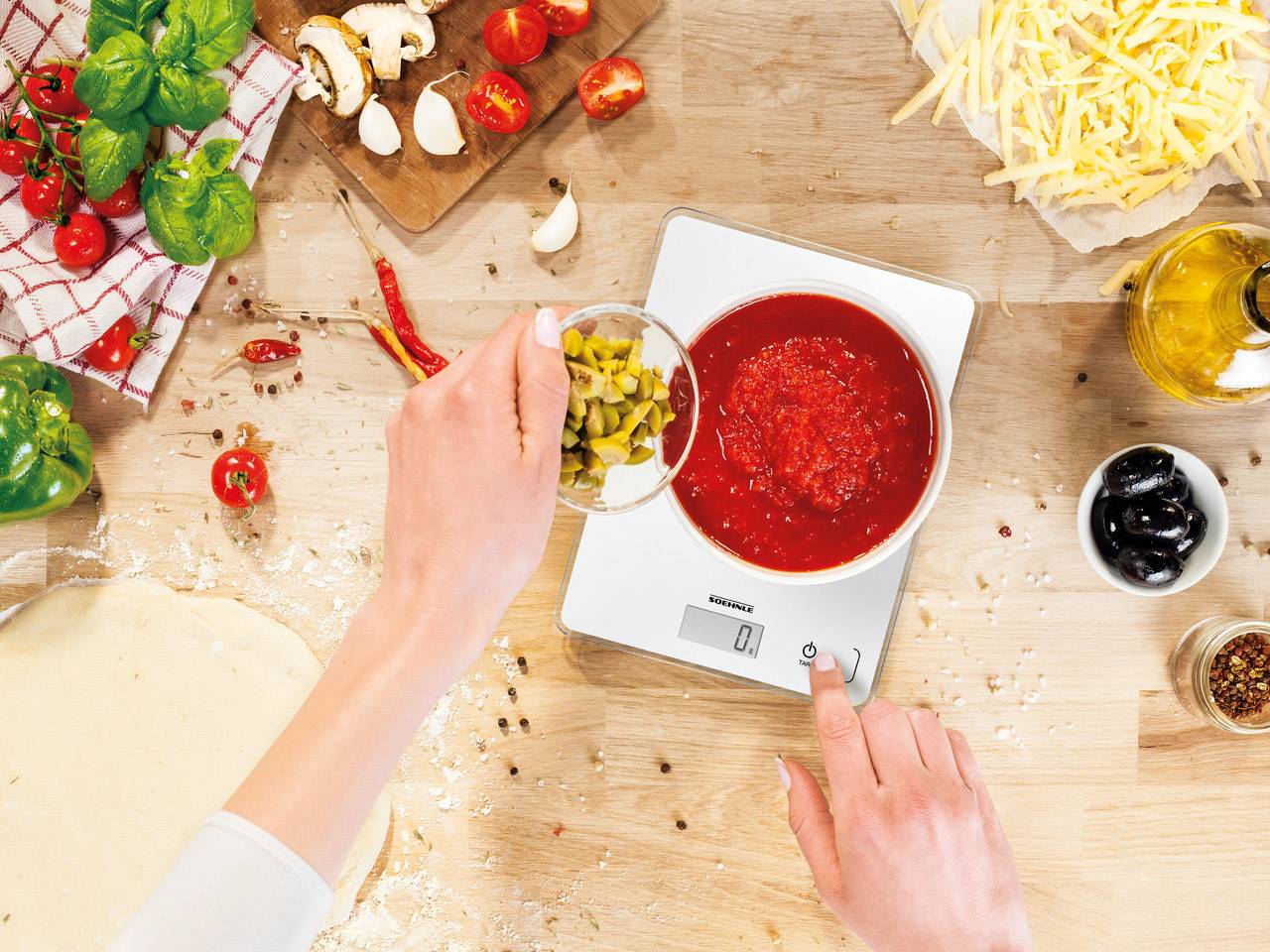 Osoba váží nasekané olivy na digitální kuchyňské váze vedle mísy s rajčatovou omáčkou, obklopena ingrediencemi na pizzu.
