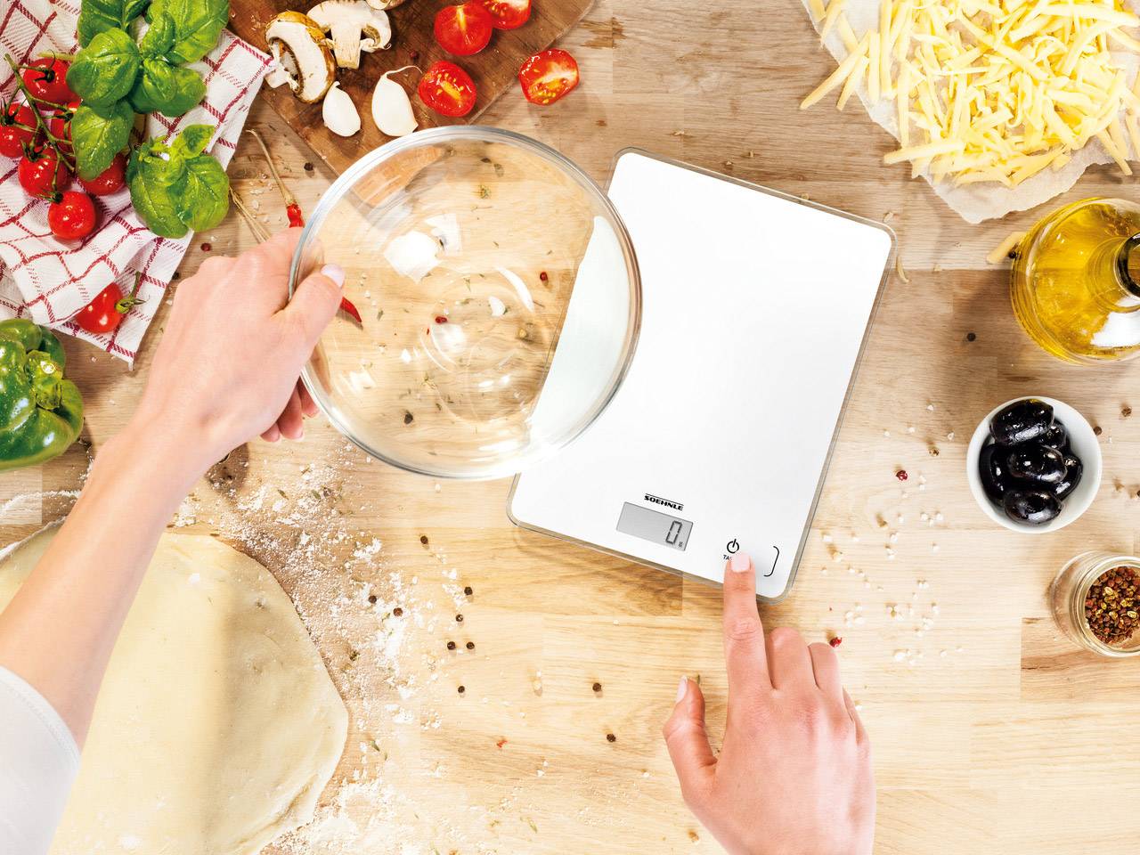 Osoba váží skleněnou mísu na digitální kuchyňské váze, obklopena ingrediencemi jako bazalka, rajčata, olivy a sýr.