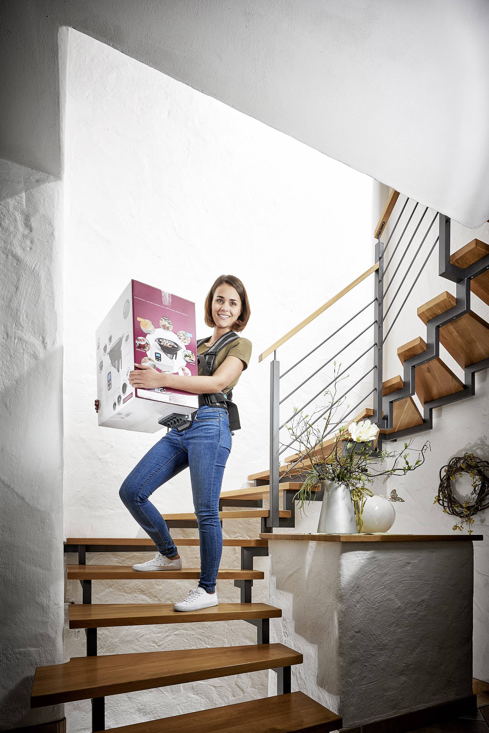 Žena nese velkou krabici dolů po schodech a usmívá se. Moderní prostředí s dřevěnými schody a rostlinnými dekoracemi v pozadí.