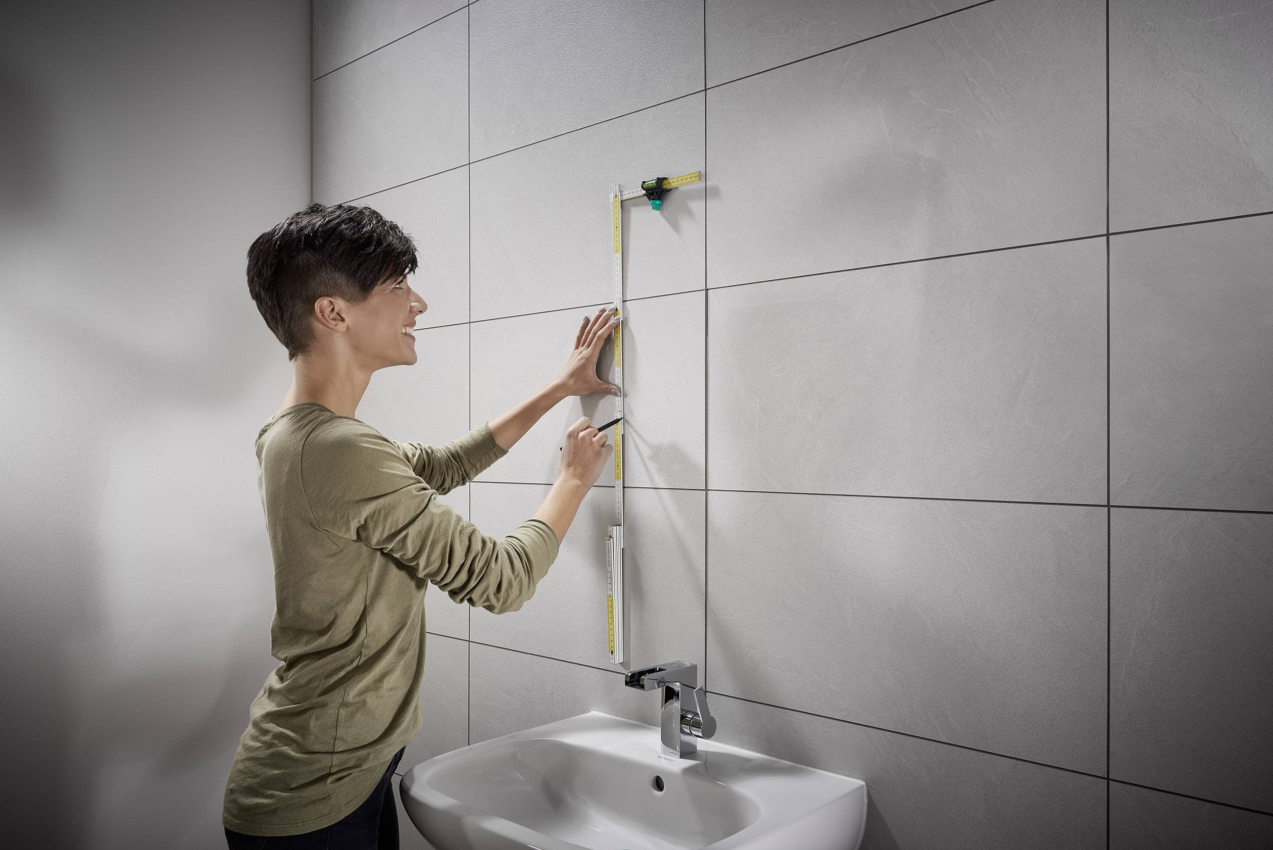 Jedna osoba měří zeď nad umyvadlem pomocí pravítka a vodní váhy.