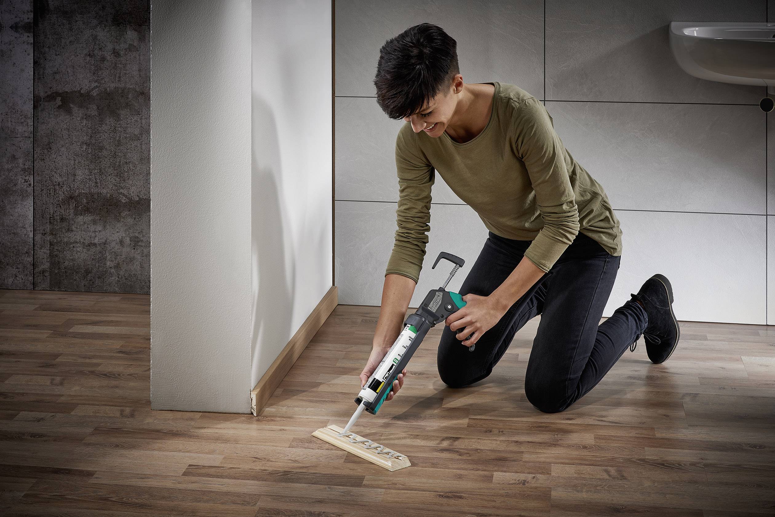 Osoba oblečená v neformálním oblečení klečí na podlaze a používá pistoli na tmely, aby upevnila dřevěnou lištu na stěnu.