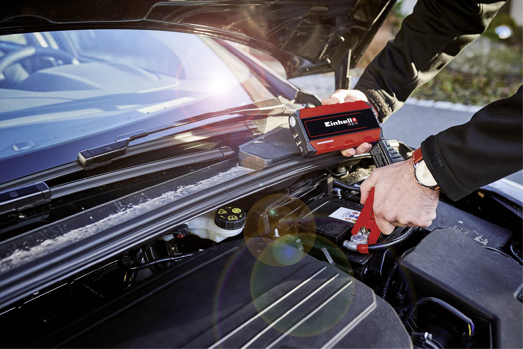 Osoba montuje autobaterii pomocí přístroje značky Einhell v motorovém prostoru automobilu.