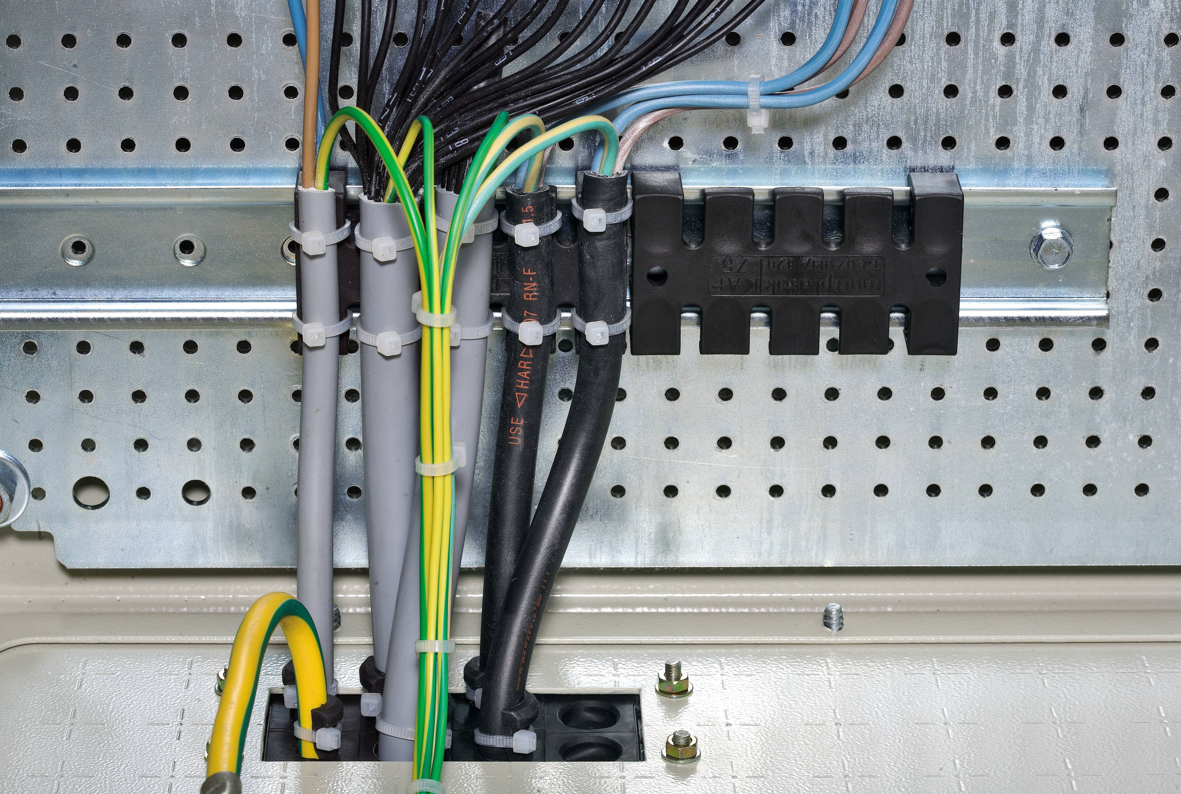 Elektrický rozvaděč s propojenými kabely v černé a šedé barvě, uzemněný žluto-zeleným kabelem, umístěný před kovovou zadní stěnou.