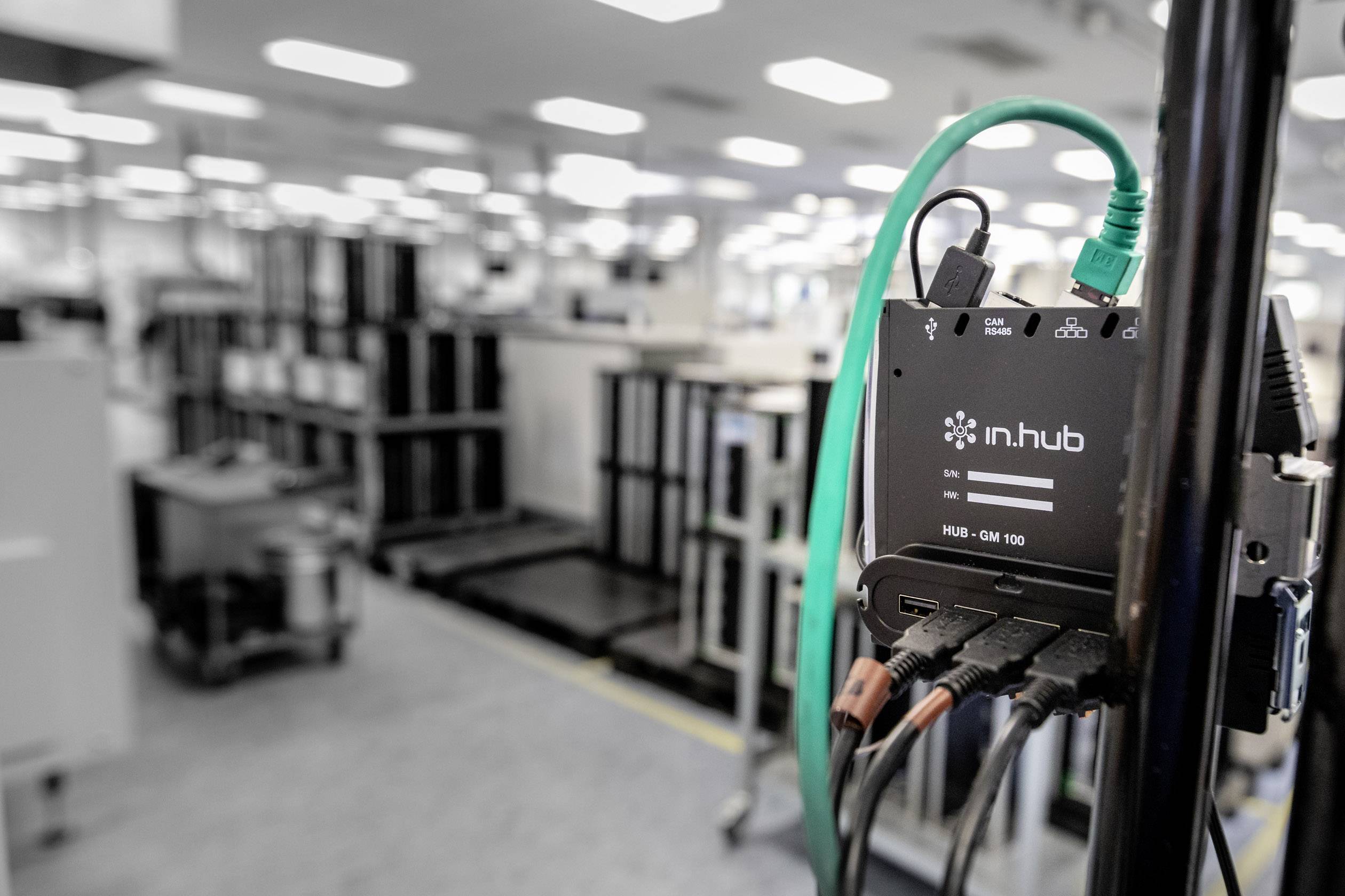 Černý hub se zelenými a černými kabely propojuje zařízení v průmyslovém prostředí. V pozadí jsou rozostřené stroje.