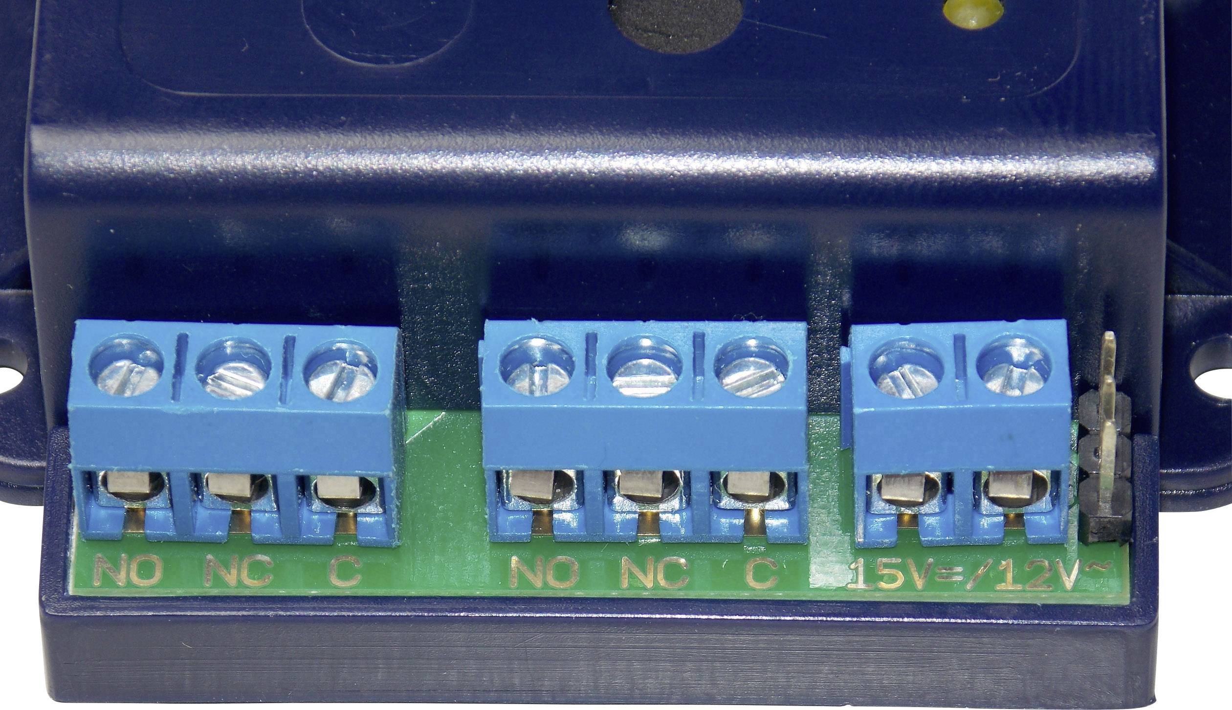 Detailní záběr modrého elektrického svorkovnicového bloku na zelené desce plošných spojů, s popisky 'NO', 'NC', 'C' a údaji o napětí.