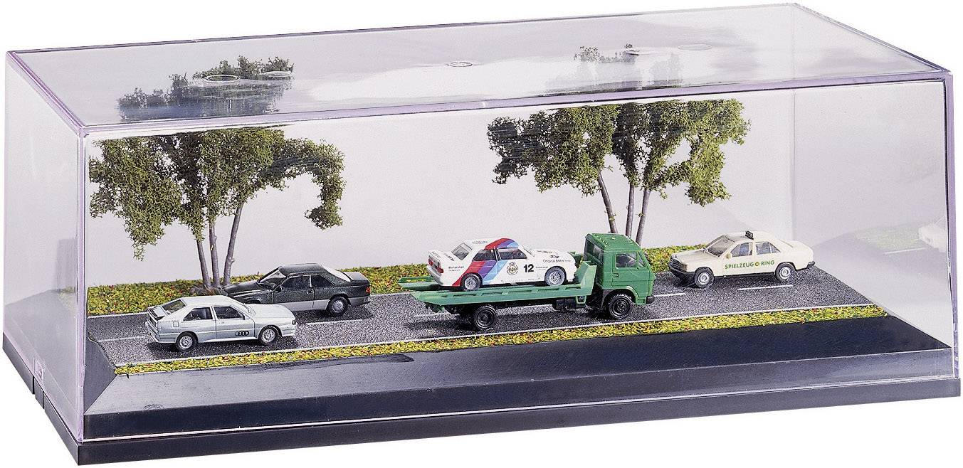 Modelový nákladní automobil přepravuje policejní vůz po silnici lemované stromy, doprovázen dvěma dalšími vozidly v průhledném displeji.