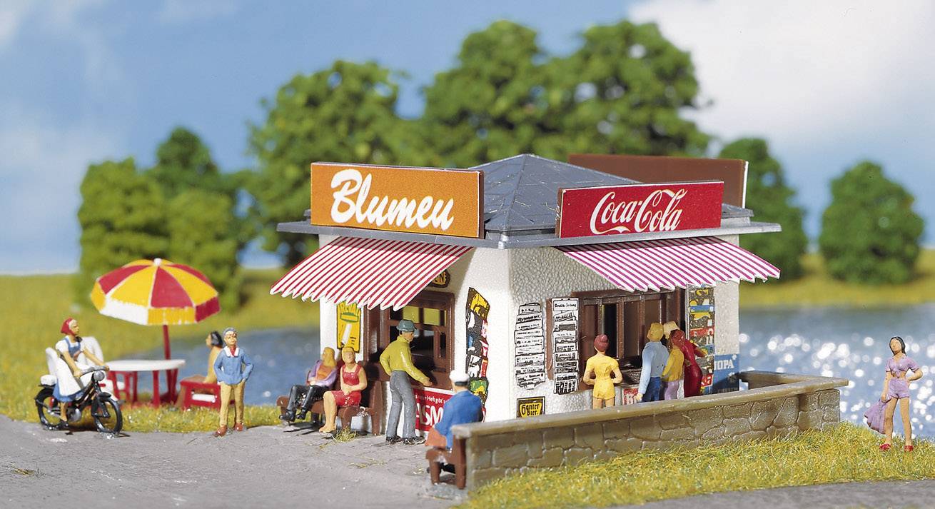 Malý stánek na břehu jezera s nápisy 'Květiny' a 'Coca-Cola', obklopený lidmi a stromy pod modrým nebem.