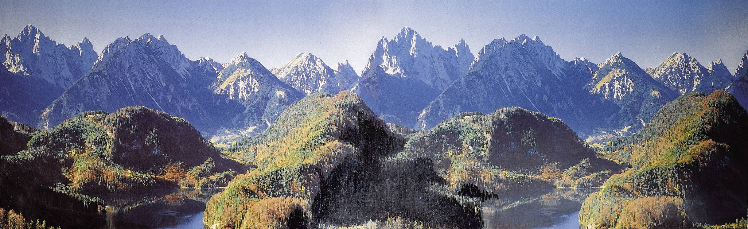 Panoramatický pohled na horské pásmo se zasněženými štíty a zalesněnými kopci v popředí pod modrou oblohou.