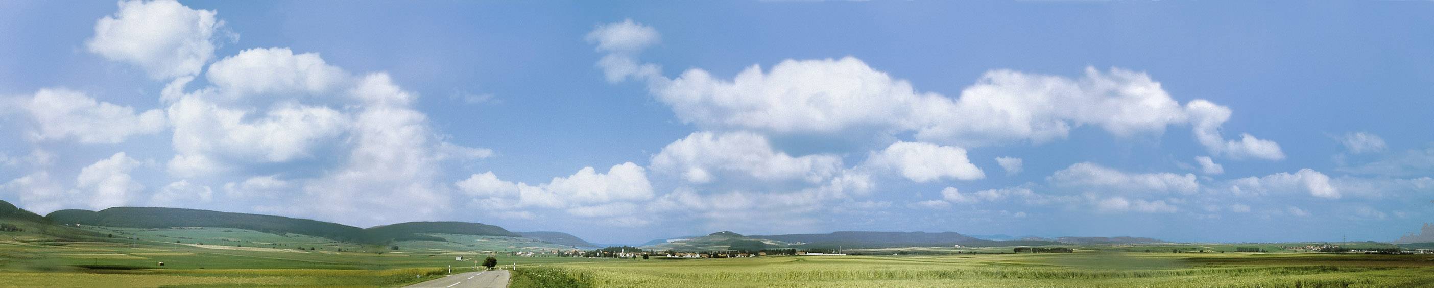 Panorama zelené krajiny s rozsáhlými poli pod modrou oblohou s bílými mraky. Na horizontu jsou viditelné kopce.