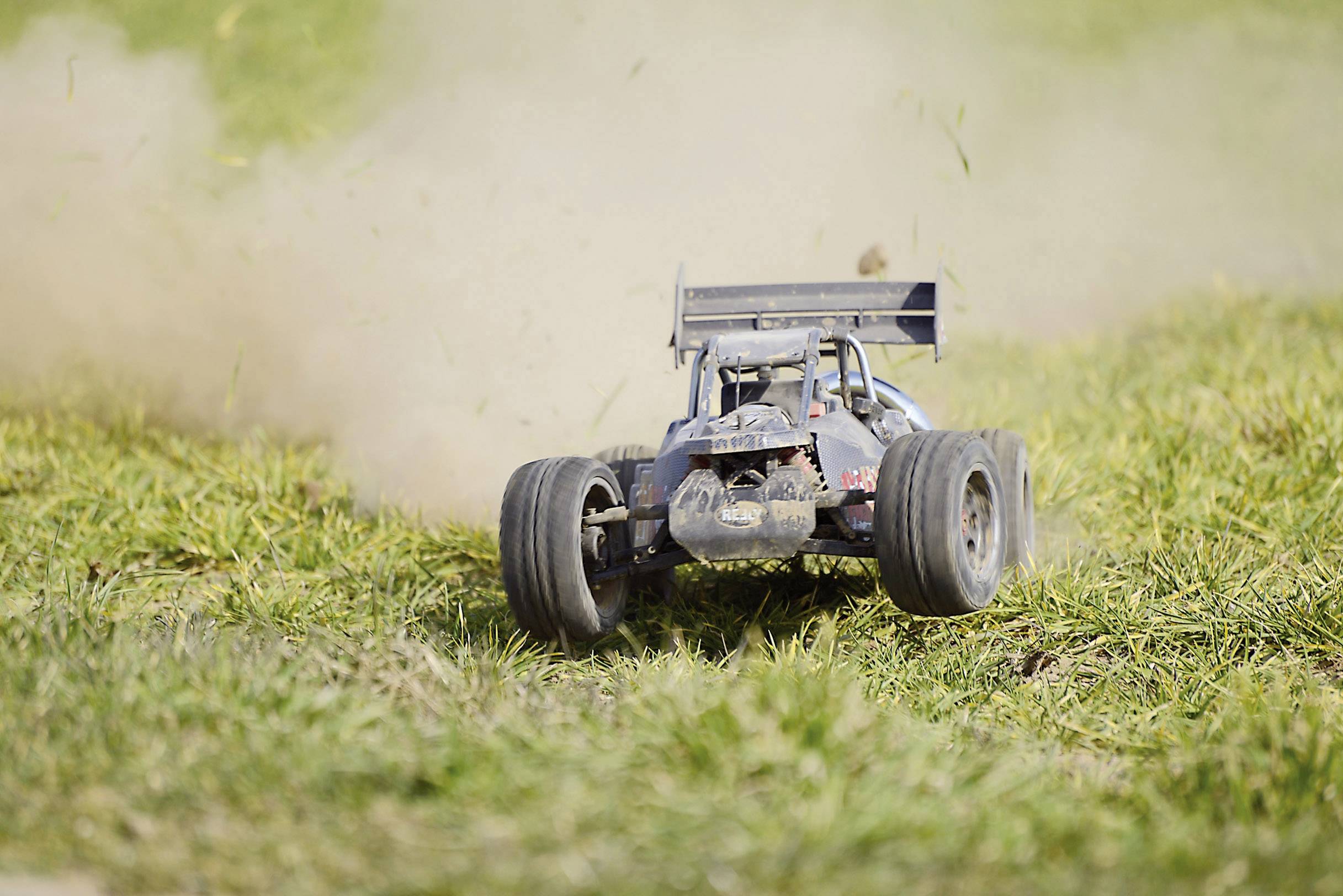 Rádiem ovládané auto jede přes trávu a zvedá prach. Závodní křídlo zlepšuje aerodynamiku. Dynamická scéna na louce.