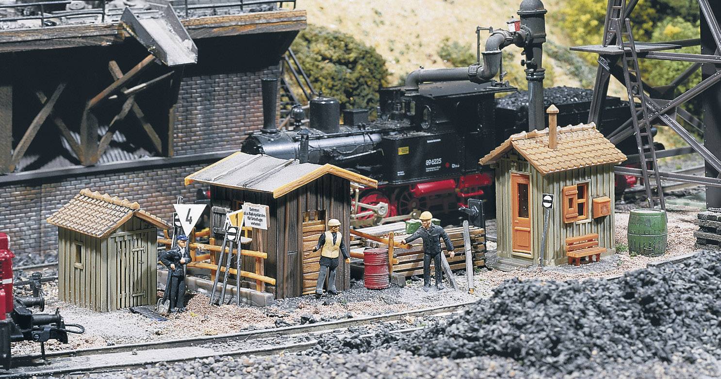Miniaturní železniční scéna s dělníky, budovami a parní lokomotivou v venkovské krajině.