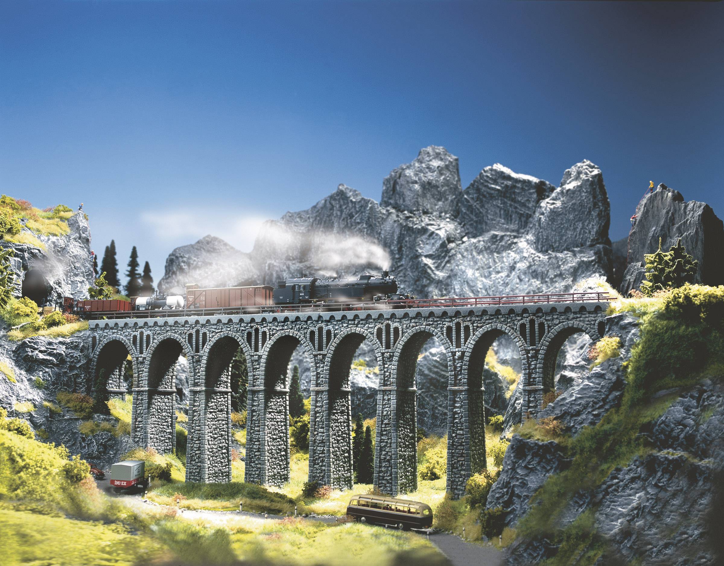 Parní lokomotiva táhne nákladní vlak po klenutém kamenném viaduktu v hornaté krajině s jasnou oblohou a zelení.