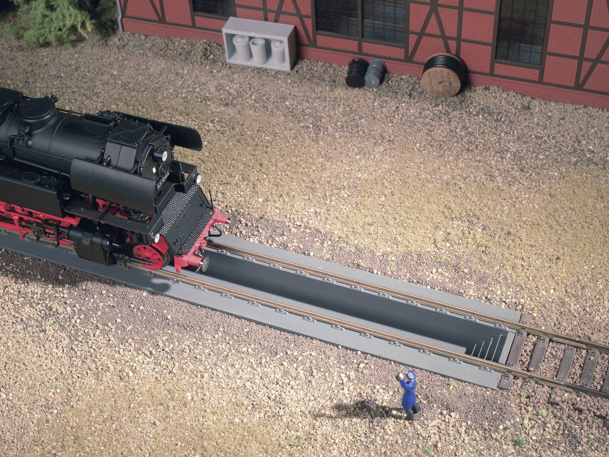 Parní lokomotiva stojí nad kolejovým servisním průchodem před budovou. Vedle ní stojí dělník v modré uniformě.