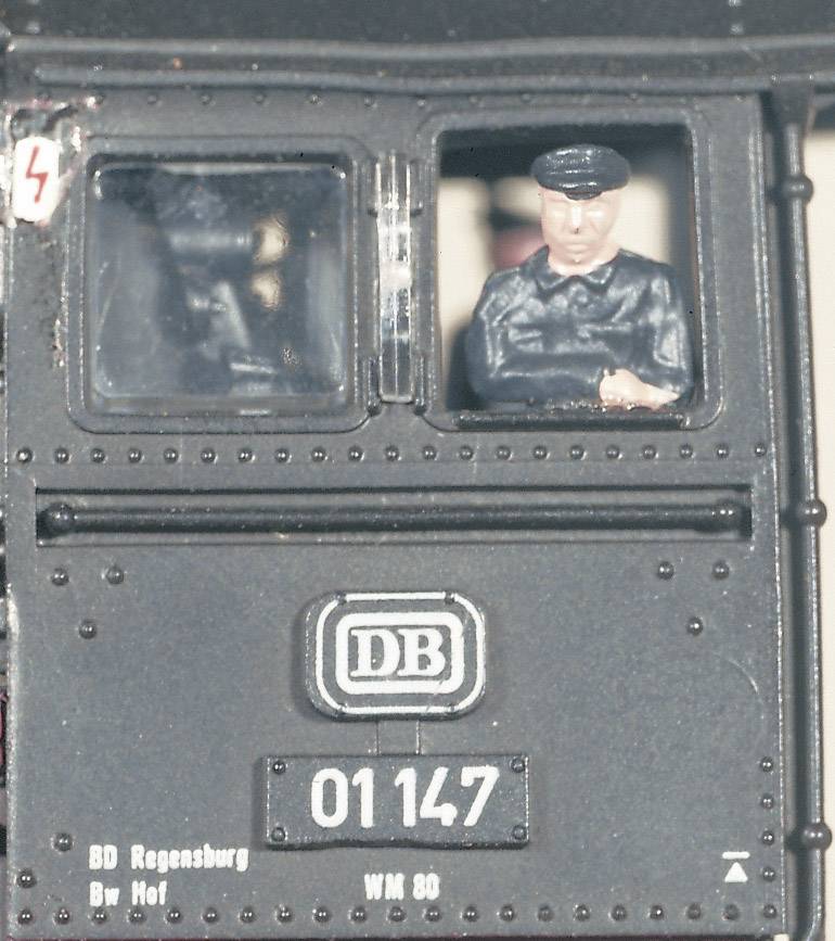 Miniaturní modelová železnice s číslem '01147' a logem 'DB'. Miniaturní figurka strojvedoucího se dívá z kabiny lokomotivy.
