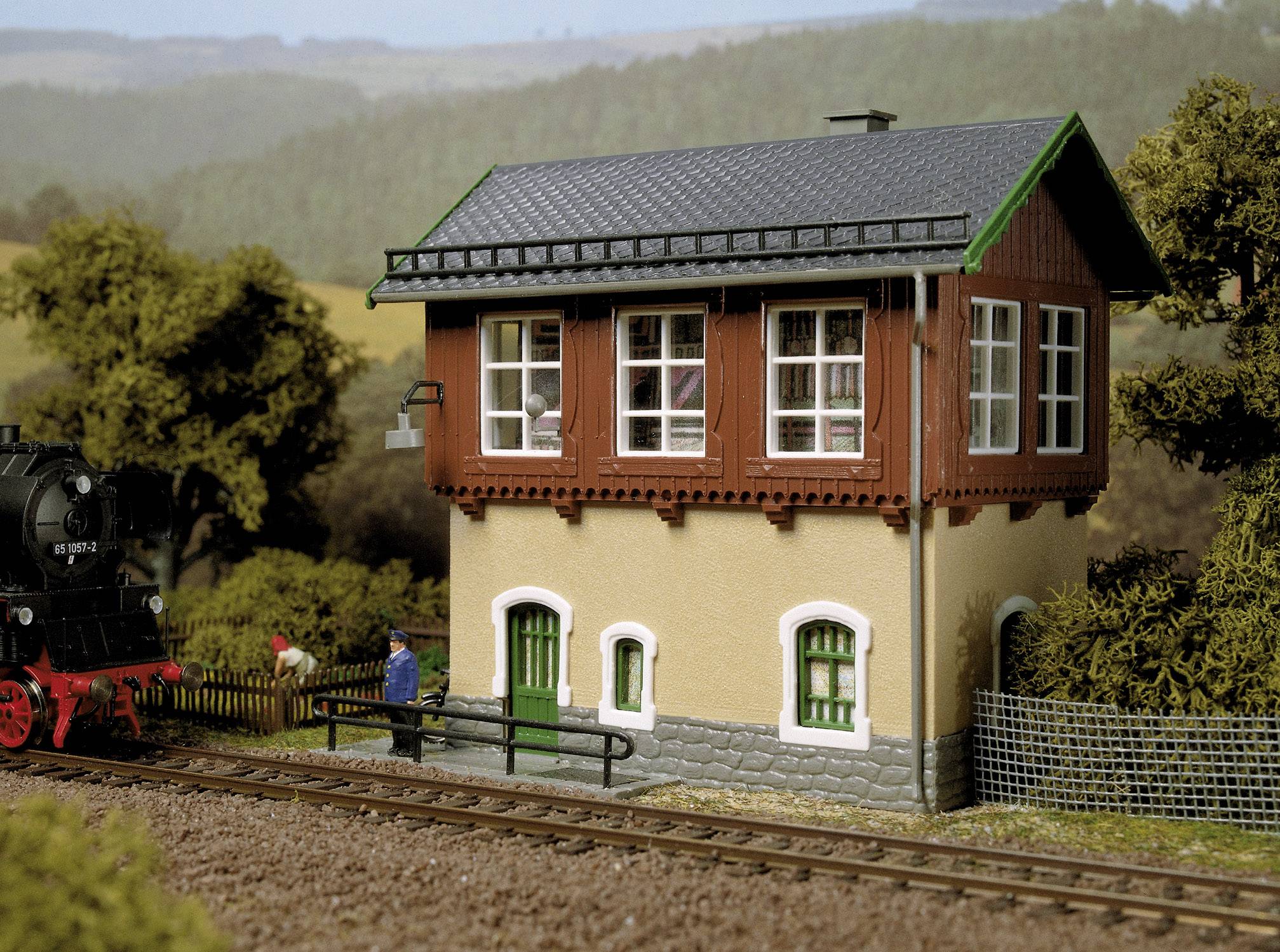 Model kolejového návěstního domku vedle železniční tratě, obklopeného stromy, v pozadí zalesněné kopce.