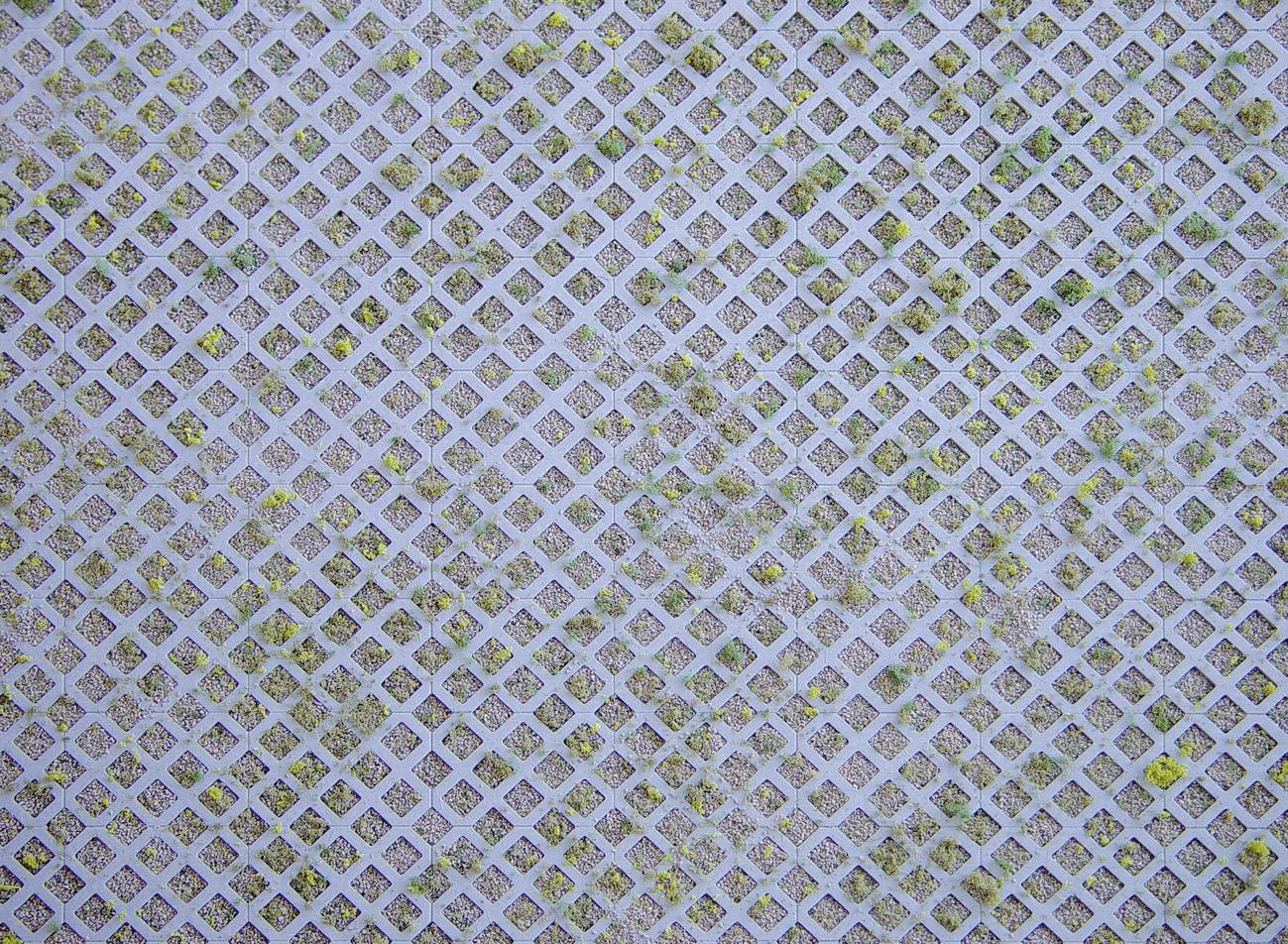 Mřížový vzor z čtvercových otvorů, převážně v šedých a hnědých tónech, s občasnými zelenými a žlutými barevnými akcenty.