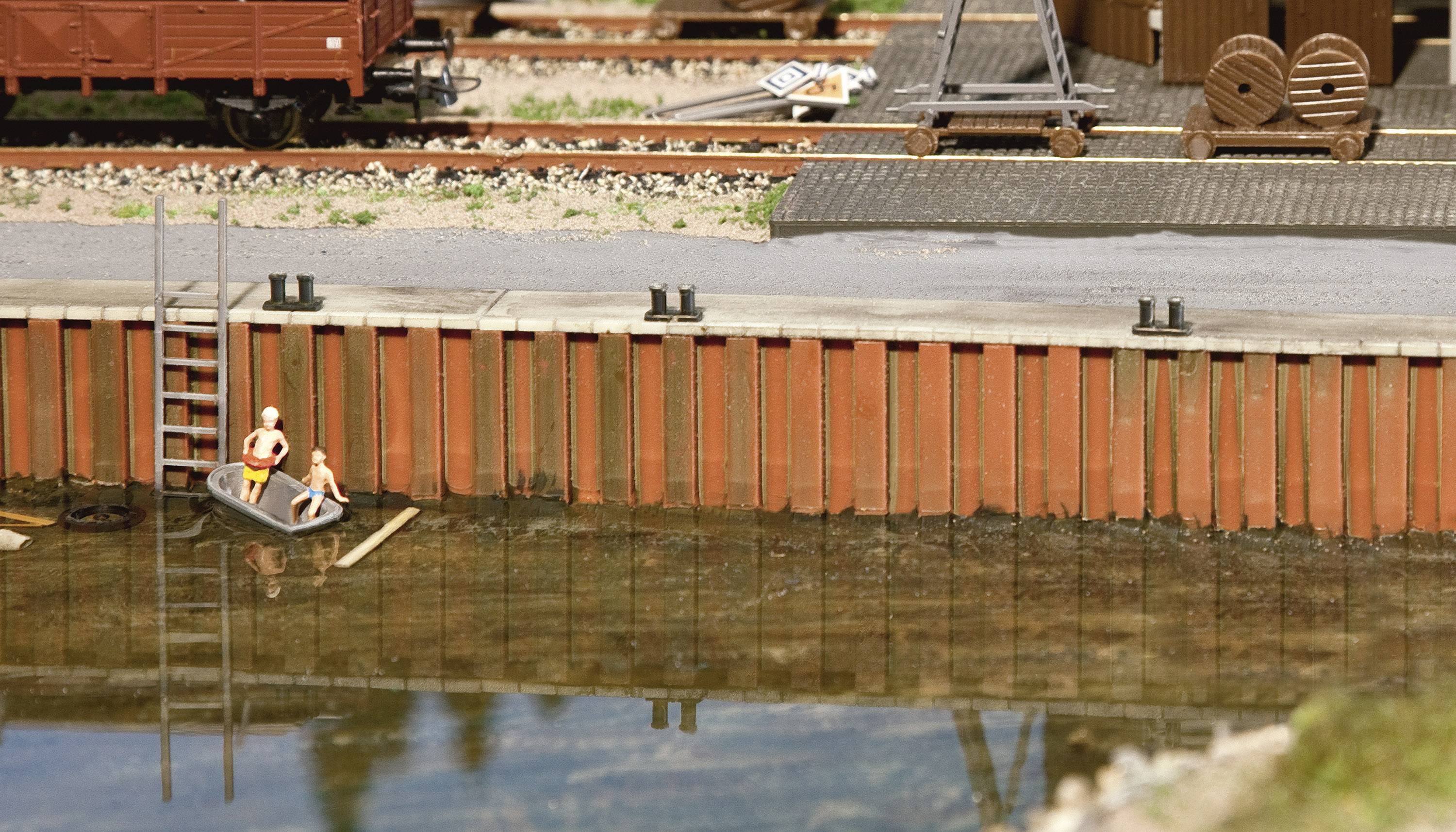 Miniaturní model zobrazuje malý přístav s lodí, ve které sedí dvě postavičky. V pozadí jsou viditelné koleje a hromady dřeva.
