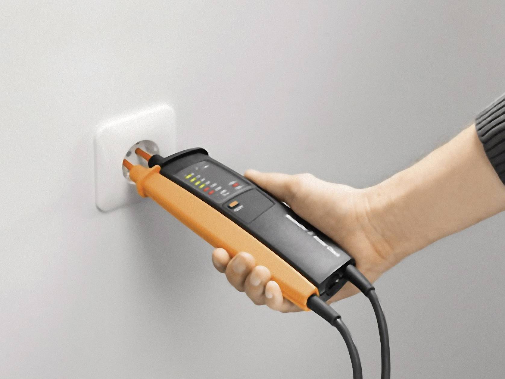 Jedna ruka drží měřicí přístroj u elektrické zásuvky pro kontrolu napětí. Přístroj zobrazuje LED indikátory pro různé úrovně napětí.