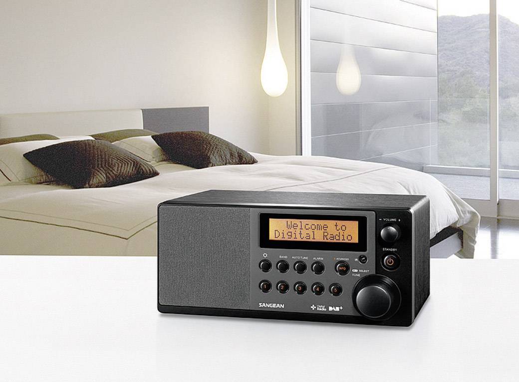 Černé digitální rádio s displejem zobrazujícím 'Vítejte v digitálním rádiu' na stole; v pozadí moderní ložnice.