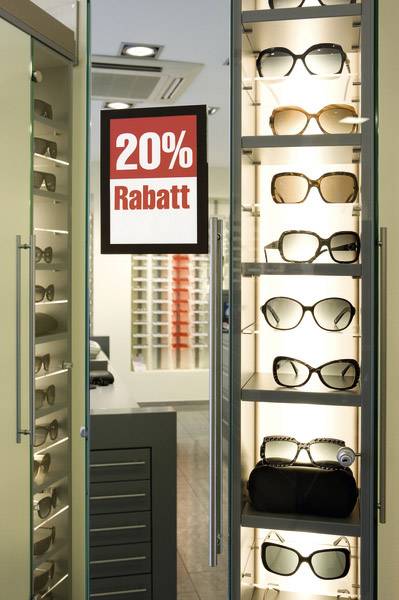 Regál s brýlemi v obchodě se slunečními brýlemi. Červená cedule ukazuje '20% sleva'.