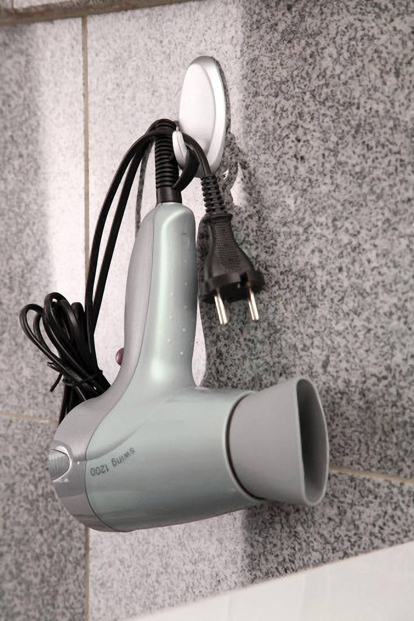 Šedý fén visí na háčku na stěně koupelny. Kabel a zástrčka jsou namotané kolem rukojeti.
