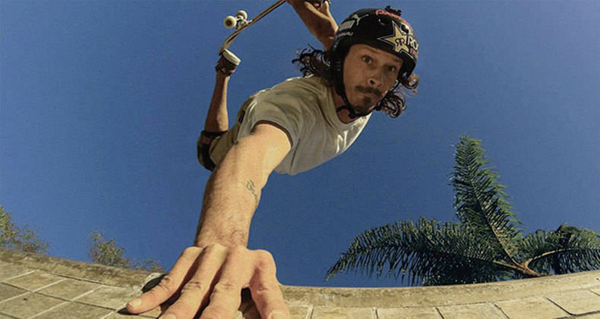 Skateboardista ve vzduchu, který se rukou dotýká rampy. Jasně modrá obloha a palma v pozadí. Akční scéna plná adrenalinu.