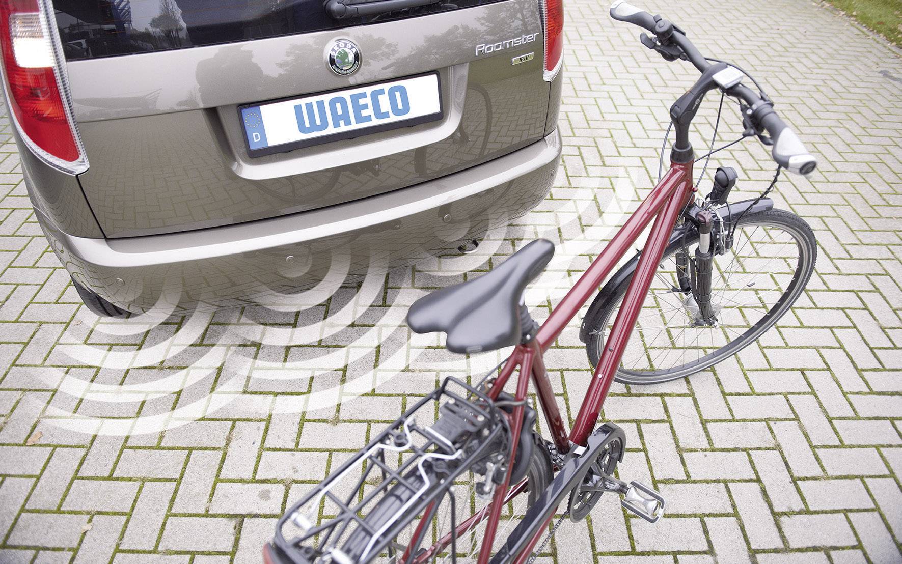 Auto parkuje pozadu na dlážděném parkovišti. Za ním stojí jízdní kolo. Vlnovky naznačují přítomnost parkovacího senzoru.