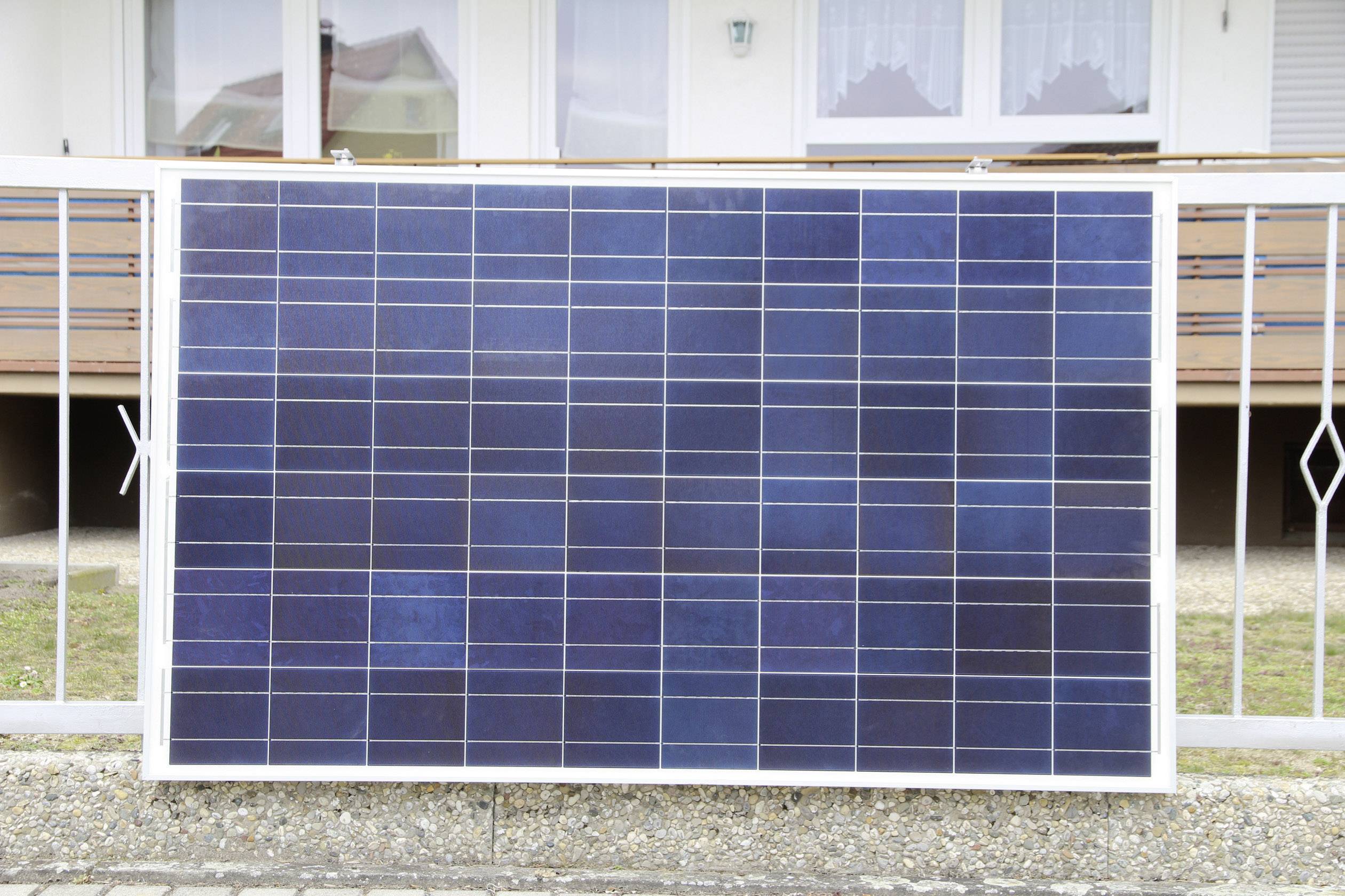Velký solární panel stojí před kovovým plotem, v pozadí je vidět dům s balkónem a okny.