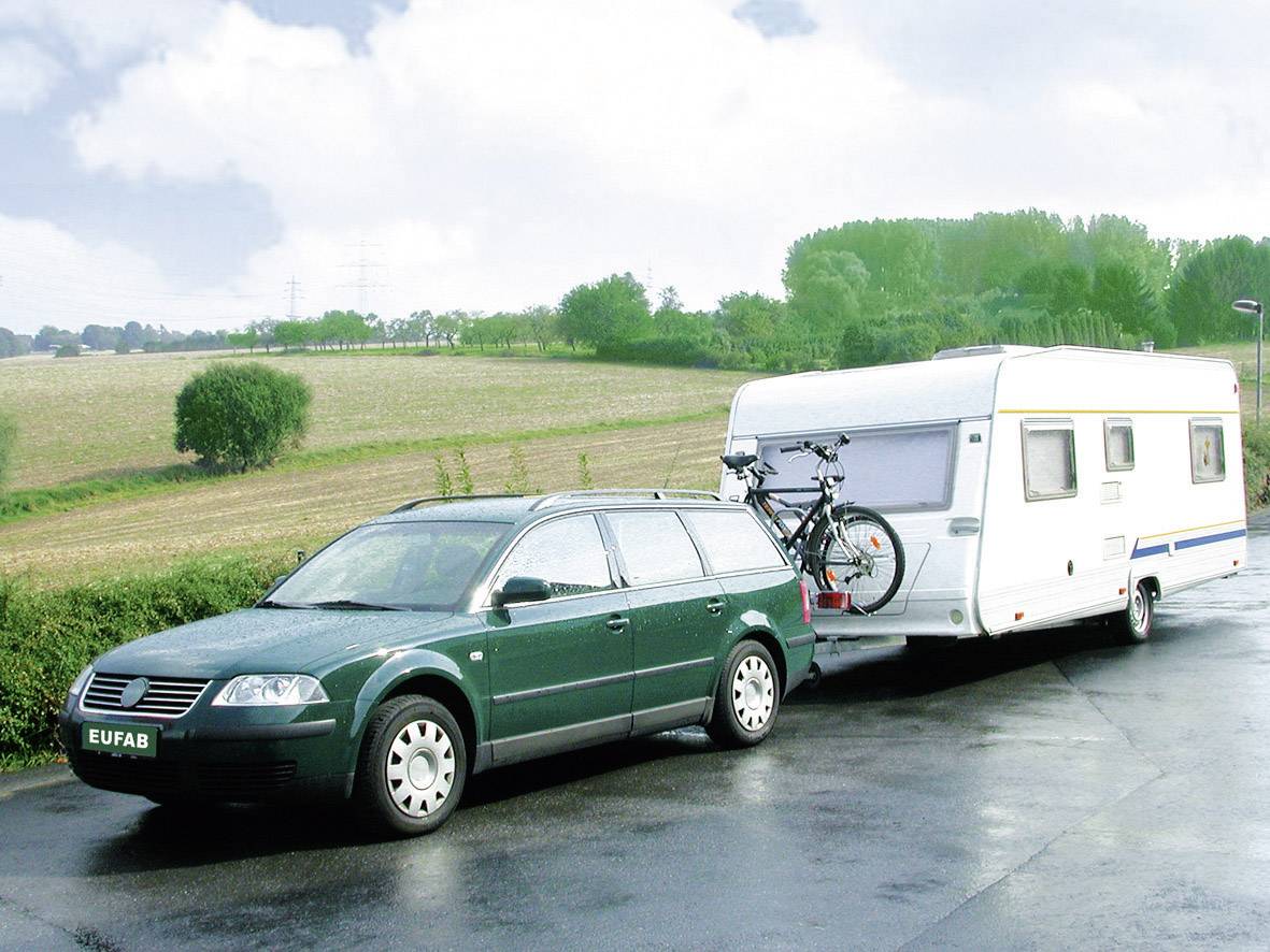 Zelené kombi táhne karavan po mokré silnici. Jízdní kolo je připevněno vzadu na autě. V pozadí jsou vidět pole a stromy.