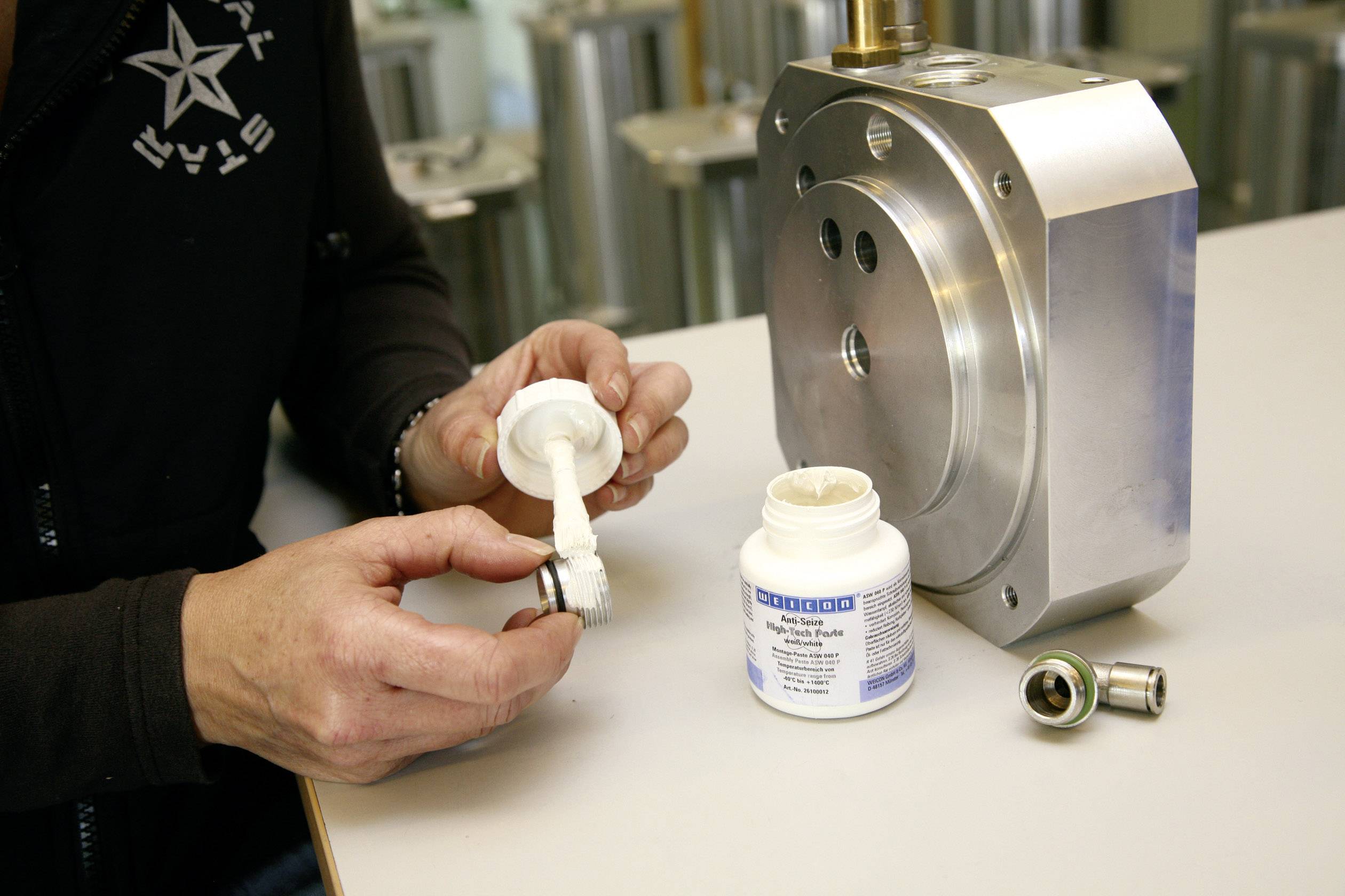 Osoba nanáší mazivo na strojní součástku. Vedle ní stojí kovová komponenta a otevřená nádoba s mazivem.
