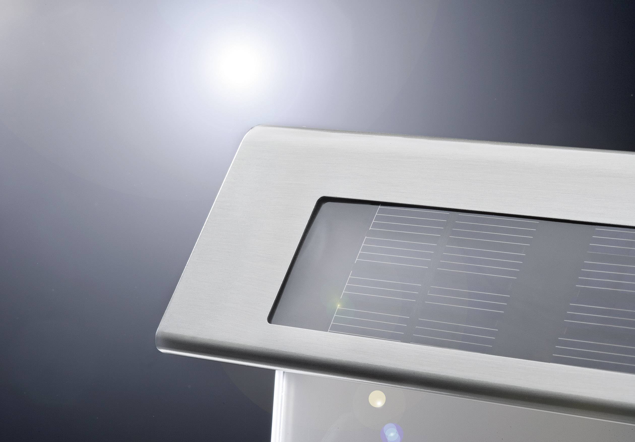 Solární panel v nerezovém rámu, snímán zblízka se slunečním svitem přímo v pozadí. Odráží světlo a zdůrazňuje moderní technologii.