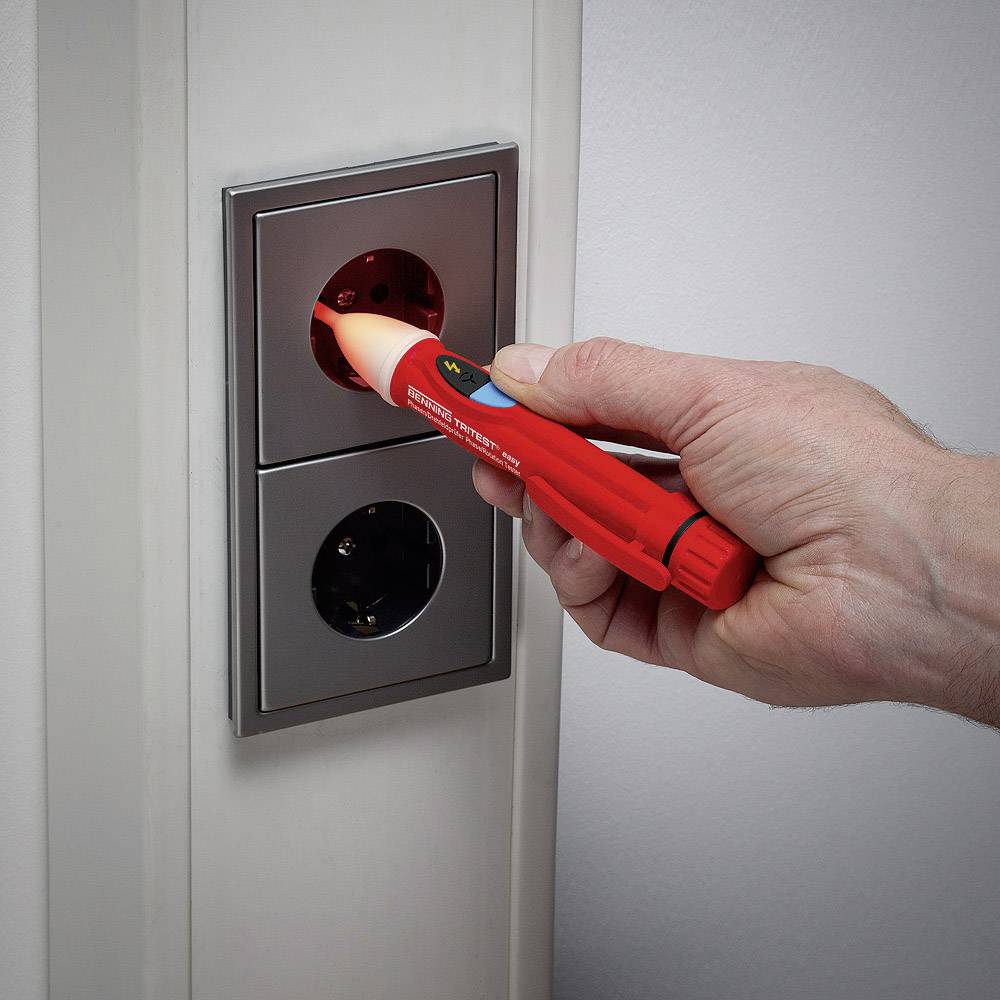 Jedna ruka přidrží zkoušečku napětí u elektrické zásuvky, aby změřila elektrické napětí. Tester zobrazuje červenou kontrolku jako výstražný signál.