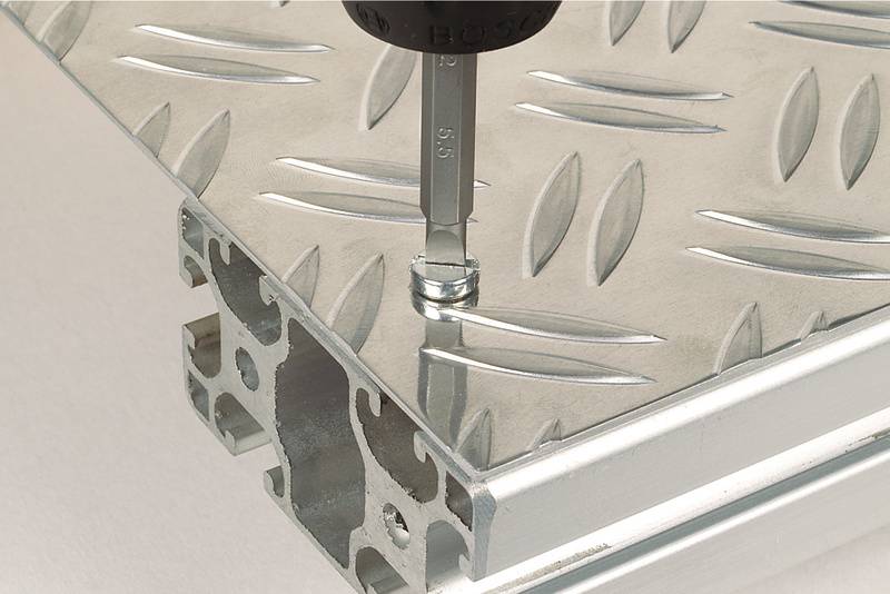 Detailní záběr šroubováku, který zašroubovává šroub do stříbrné kovové desky s protiskluzovým kosočtvercovým vzorem.