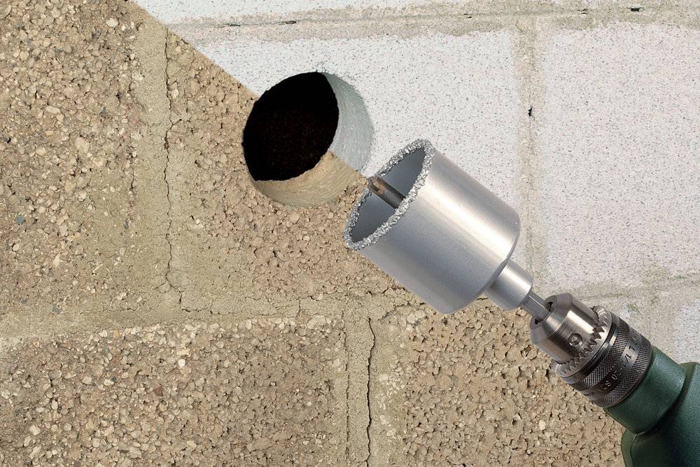 Vrtačka s vykružovací pilou vrtá kruhový otvor do betonové stěny. Vlevo je vidět nezpracovaný zdící kámen, vpravo opracovaný beton.